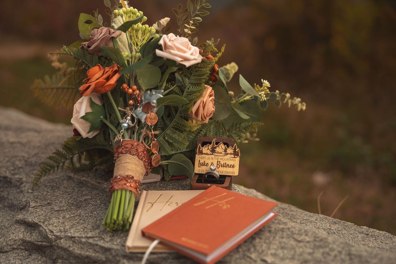 A wedding bouquet with orange and pink roses sits on a rock next to a coral-colored book.