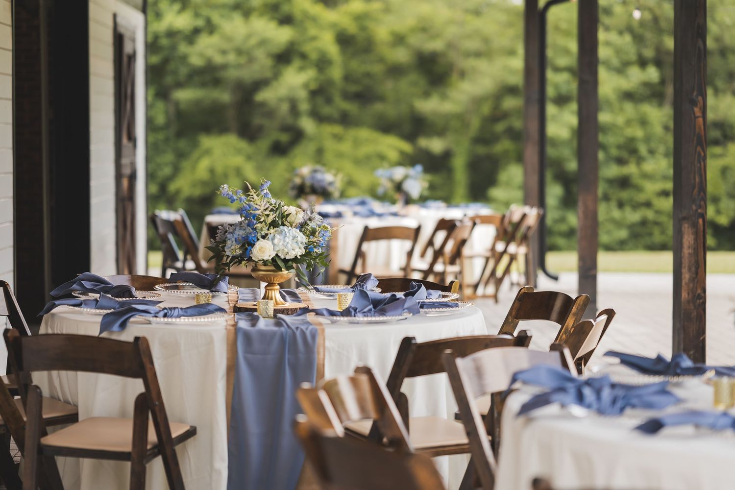 Elegant wedding reception tables set with blue linens and floral centerpieces in a glass-enclosed venue overlooking nature.