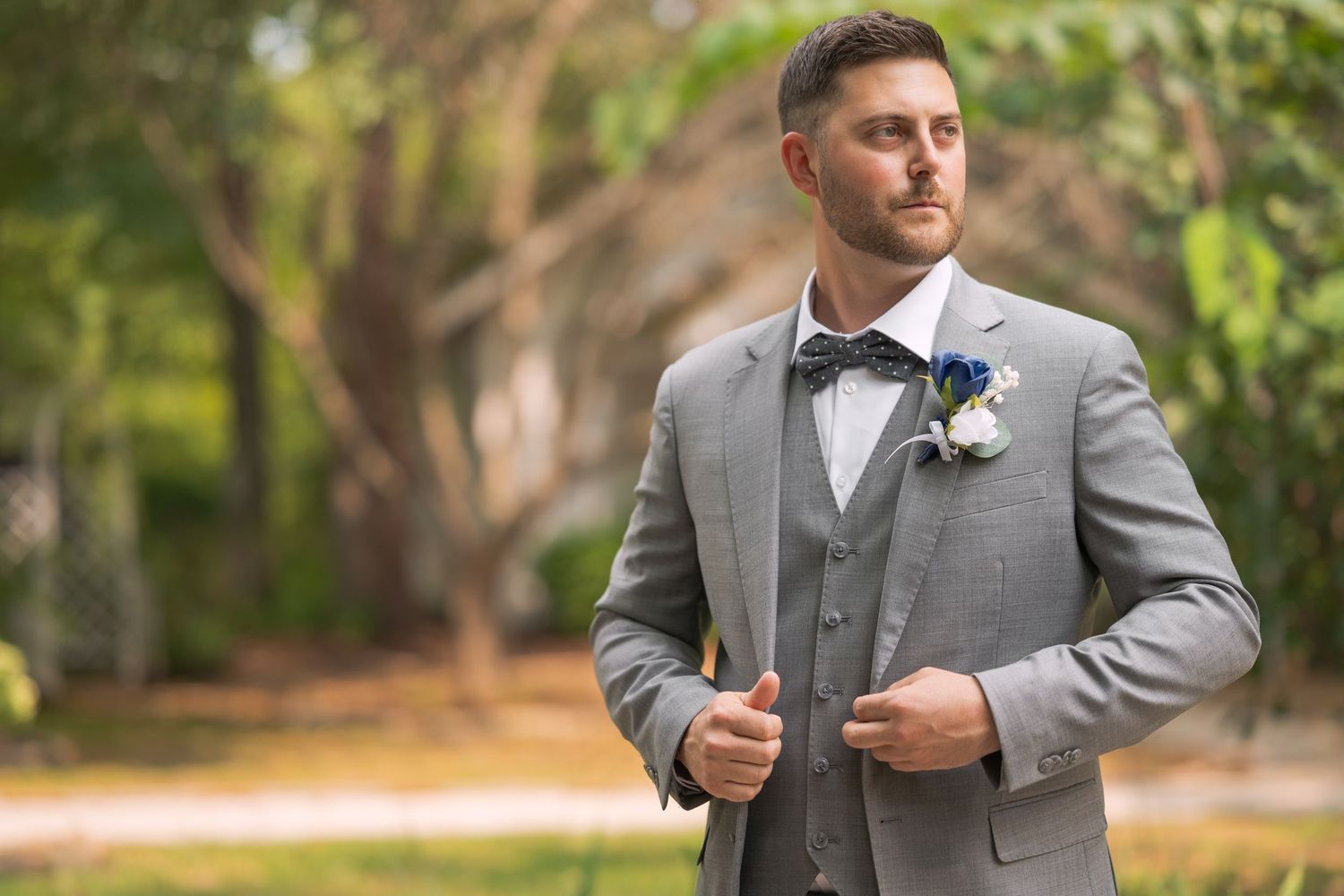 A person in a light gray suit and tie stands outdoors in a lush garden setting.