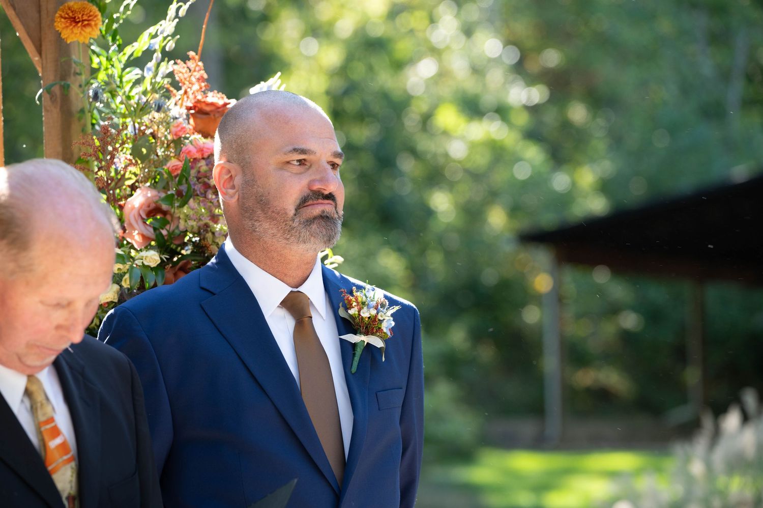 Wedding guests gather under trees at an outdoor ceremony with a wooden arch and stone water feature.