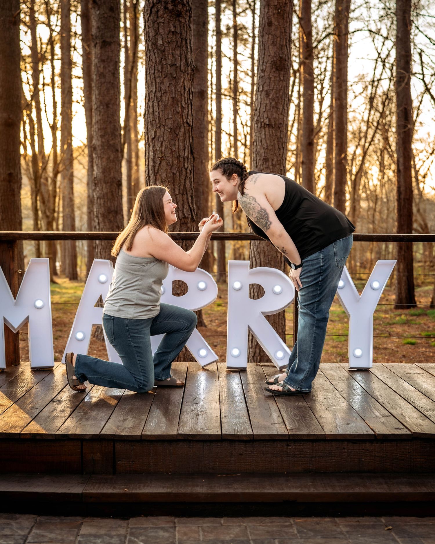 A couple kneels on a wooden deck in front of illuminated MARRY letters during a sunset marriage proposal in the woods.