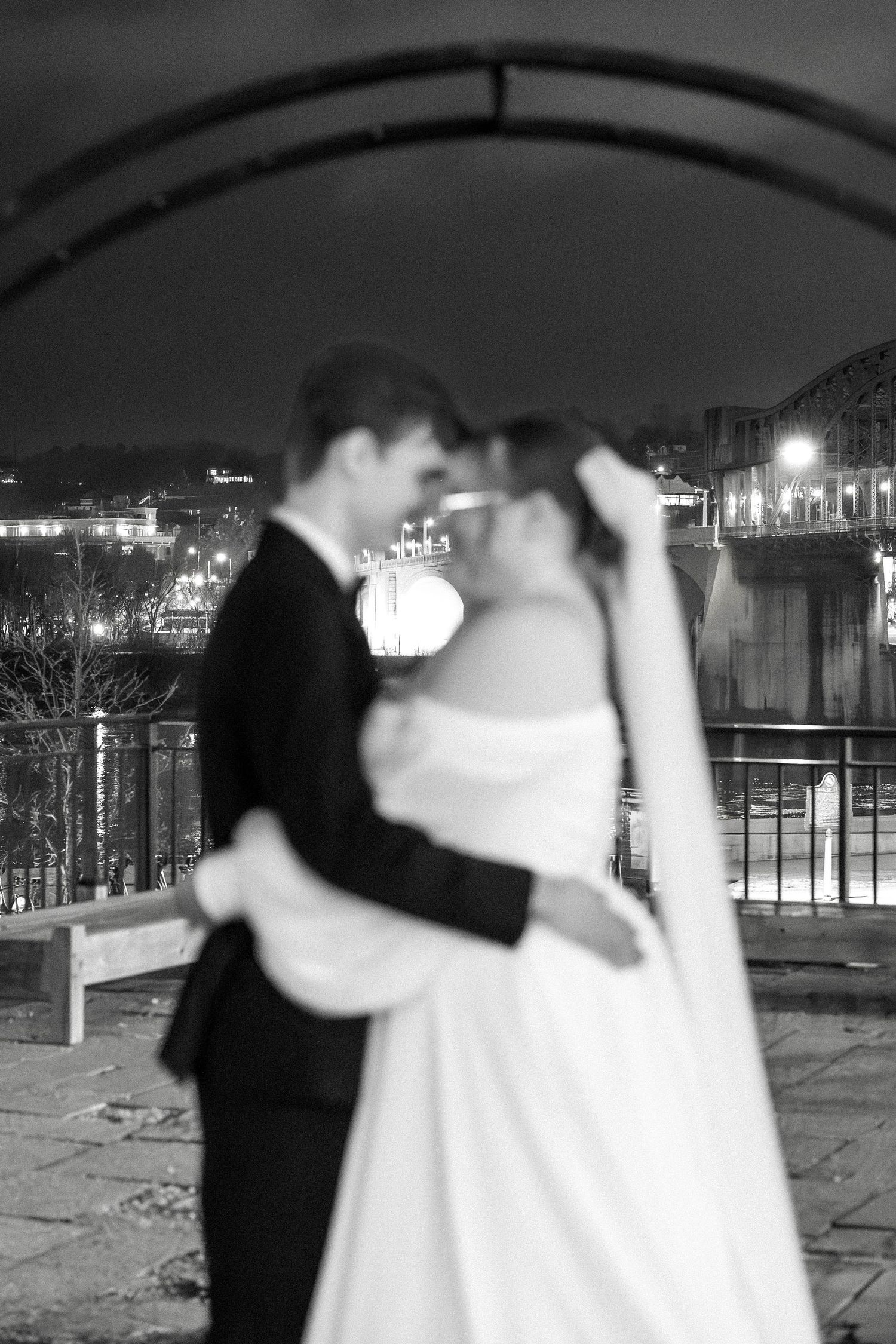 A romantic black and white nighttime portrait of newlyweds embracing on a balcony with city lights in background.