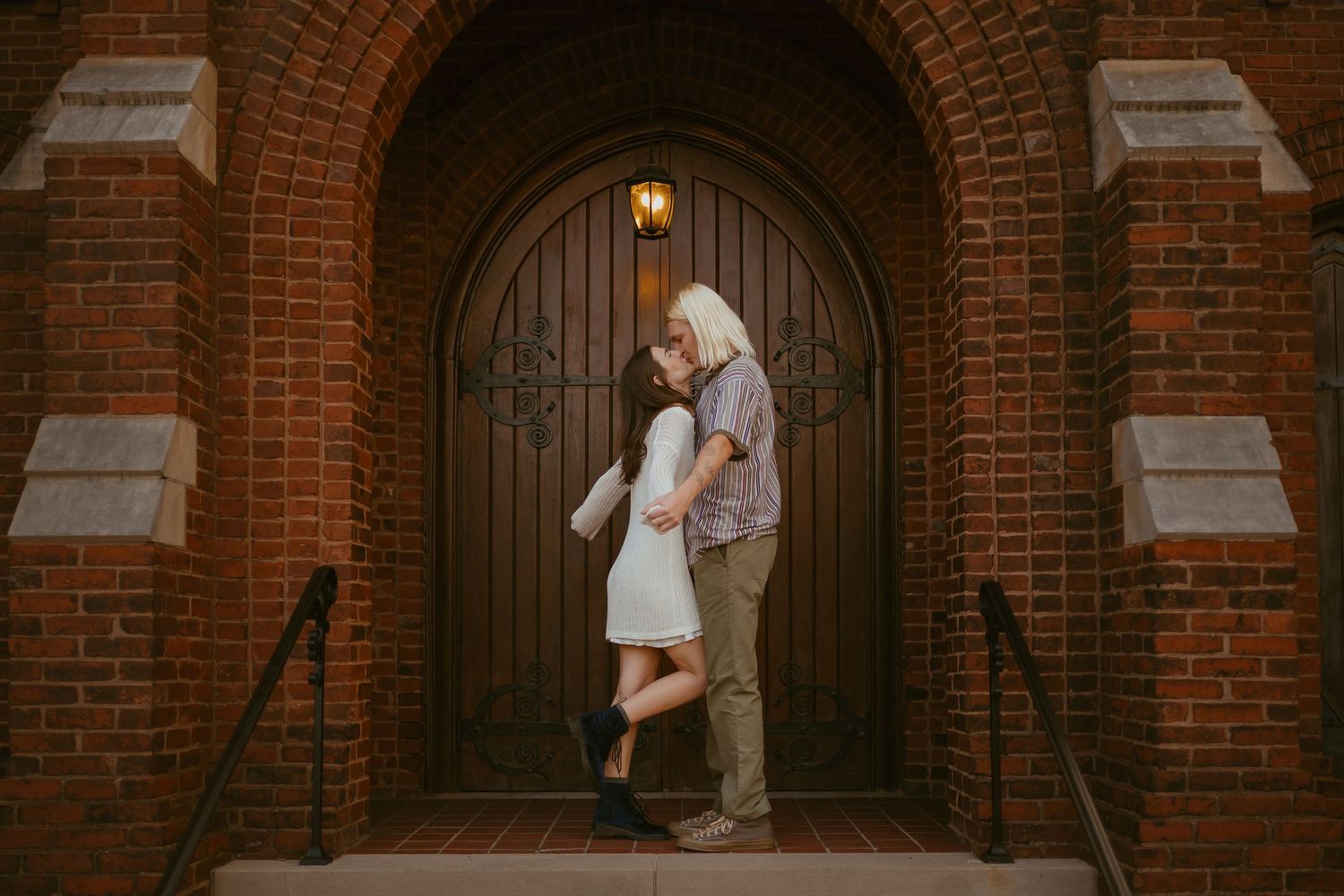A couple shares an intimate moment in front of a grand arched wooden doorway with red brick architecture.