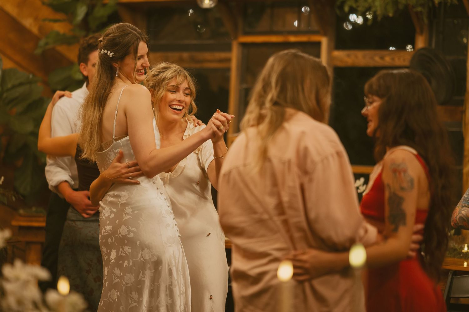 Wedding reception guests dance and celebrate in a warmly lit rustic venue with string lights overhead.