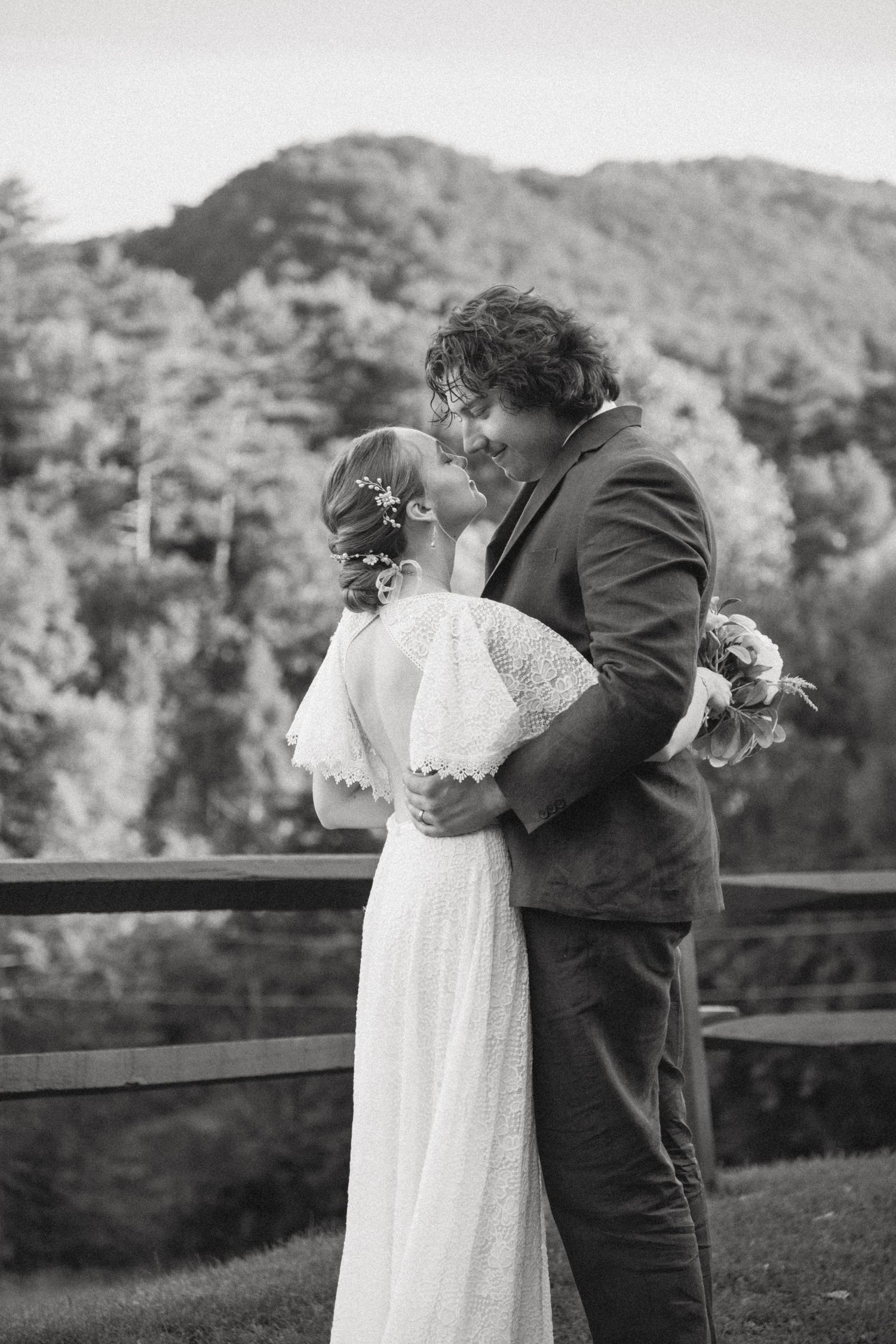 A tender black and white wedding moment captured outdoors with a mountain backdrop and rustic wooden railing.