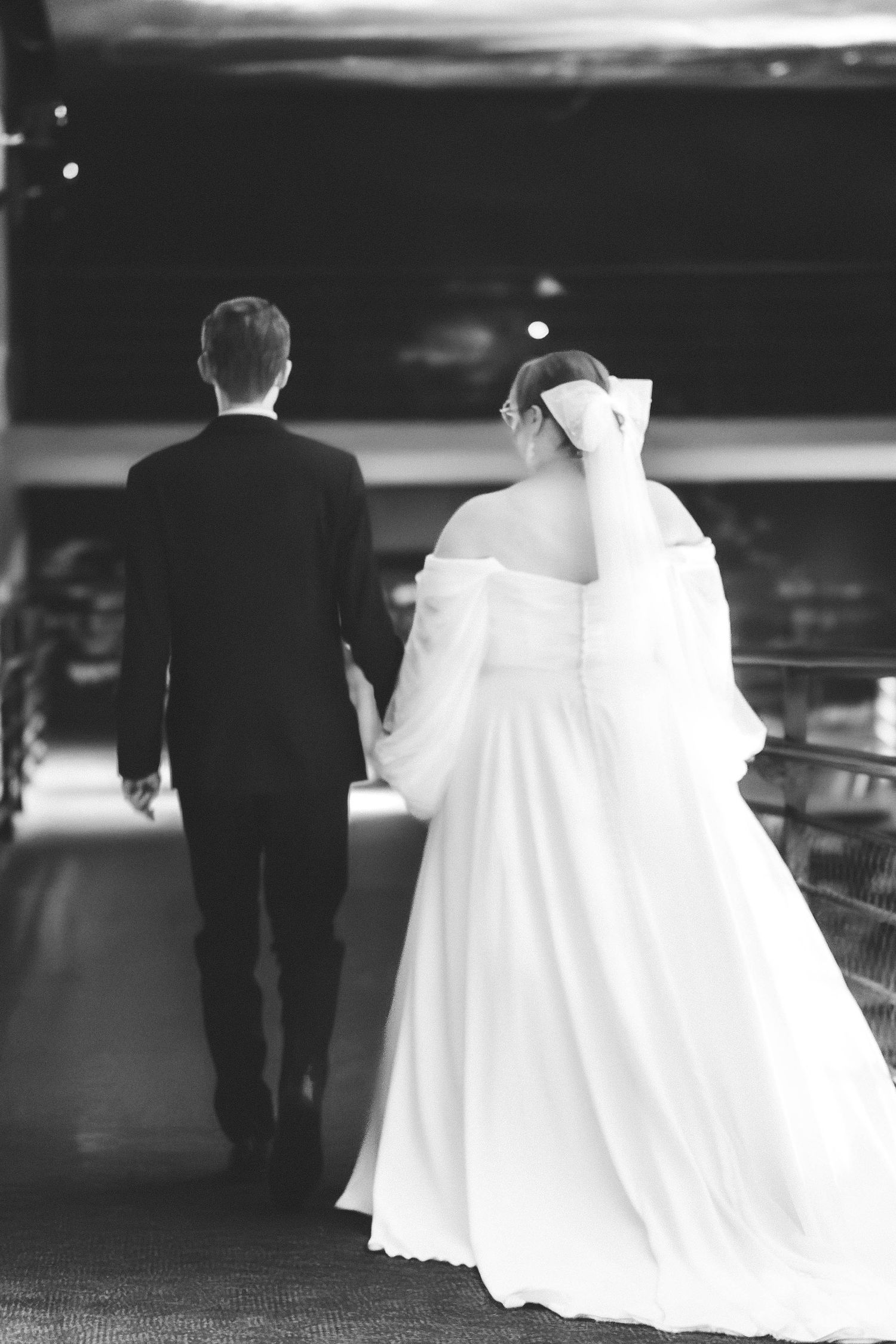 A bride in a flowing white dress and groom in black suit walk away from camera in romantic black and white photo.