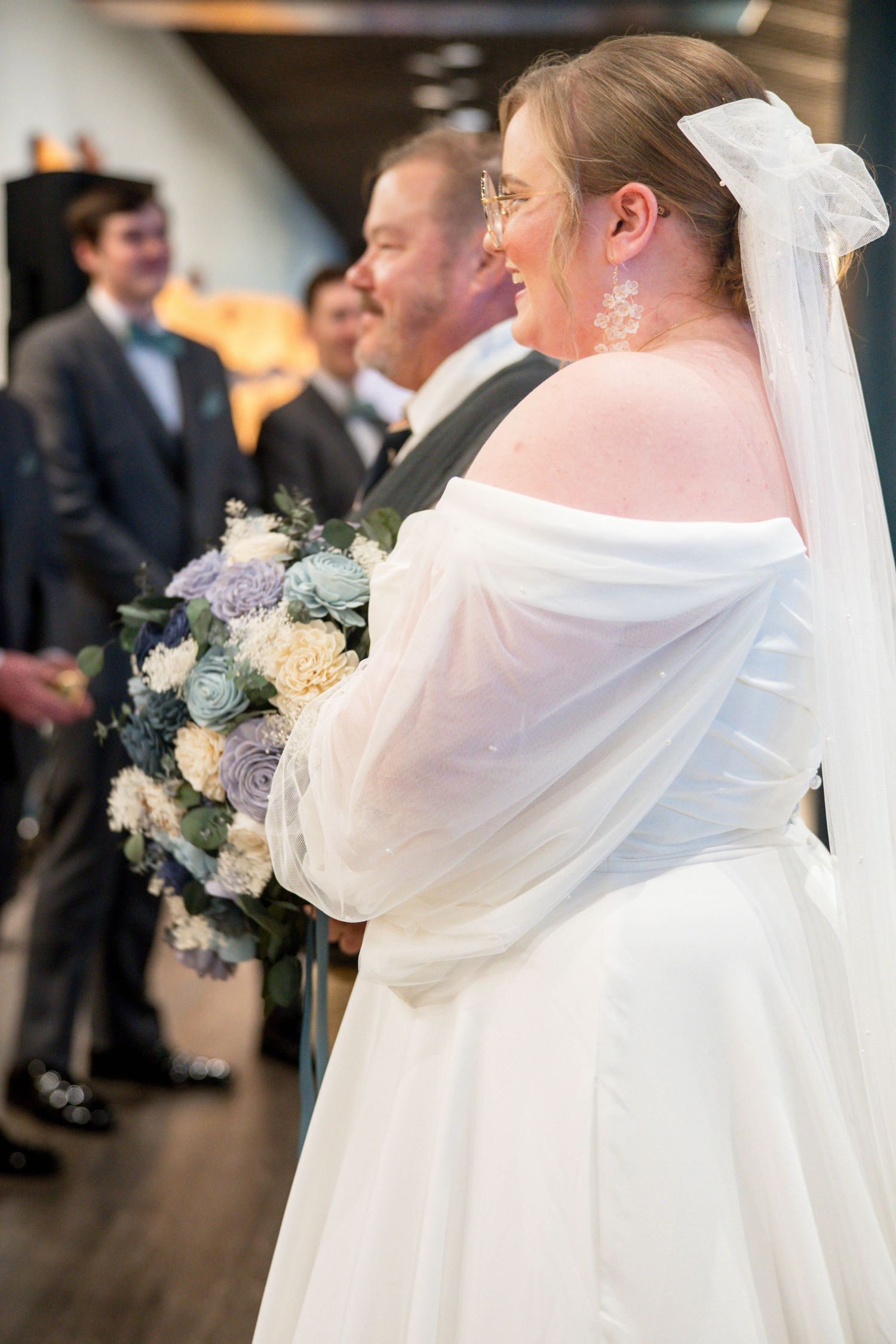 Side view of a bride in an off-shoulder white wedding dress holding a blue and white bouquet during the ceremony.