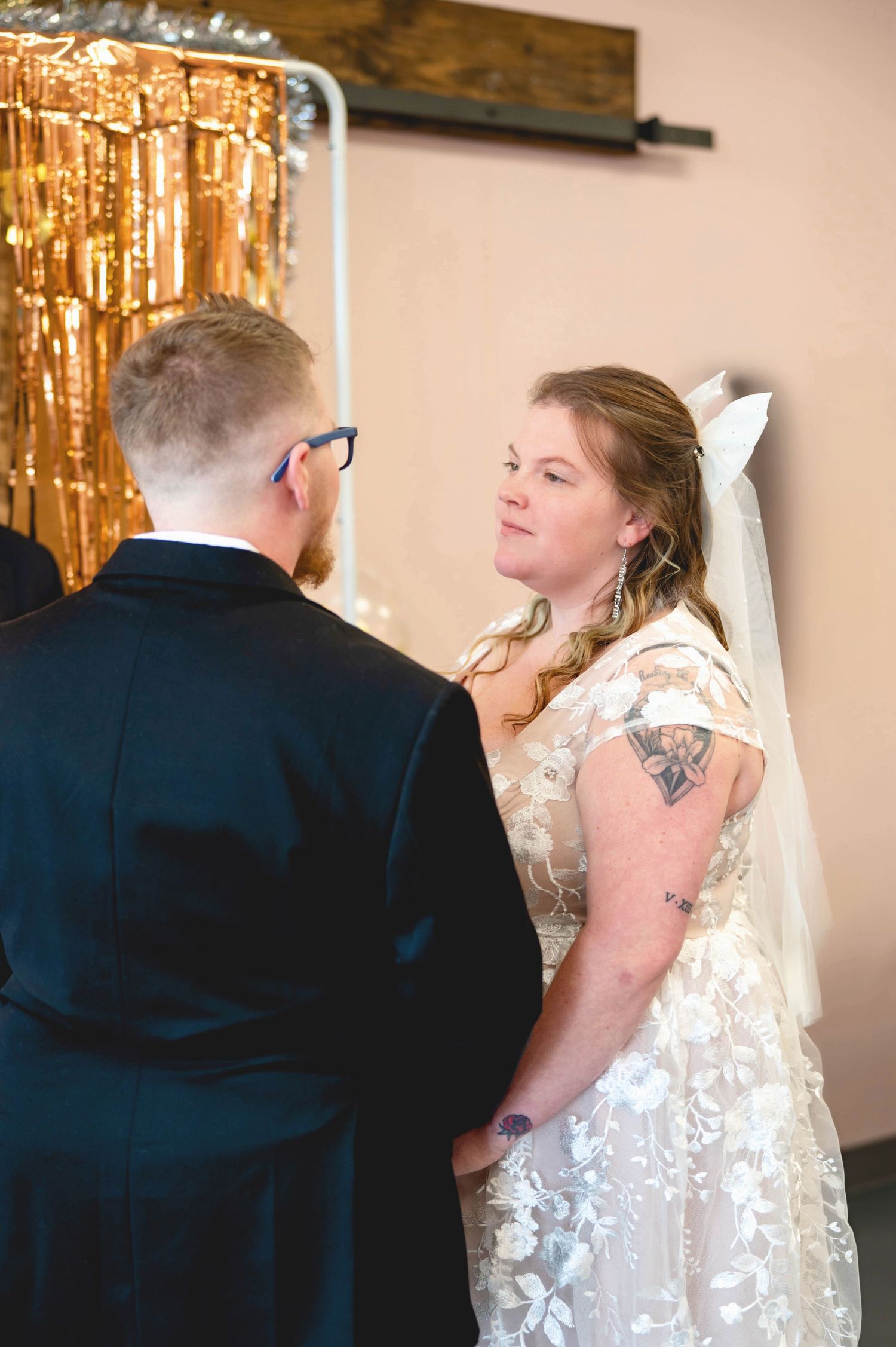 Two people in formal wedding attire stand together during ceremony with gold tinsel backdrop behind them.
