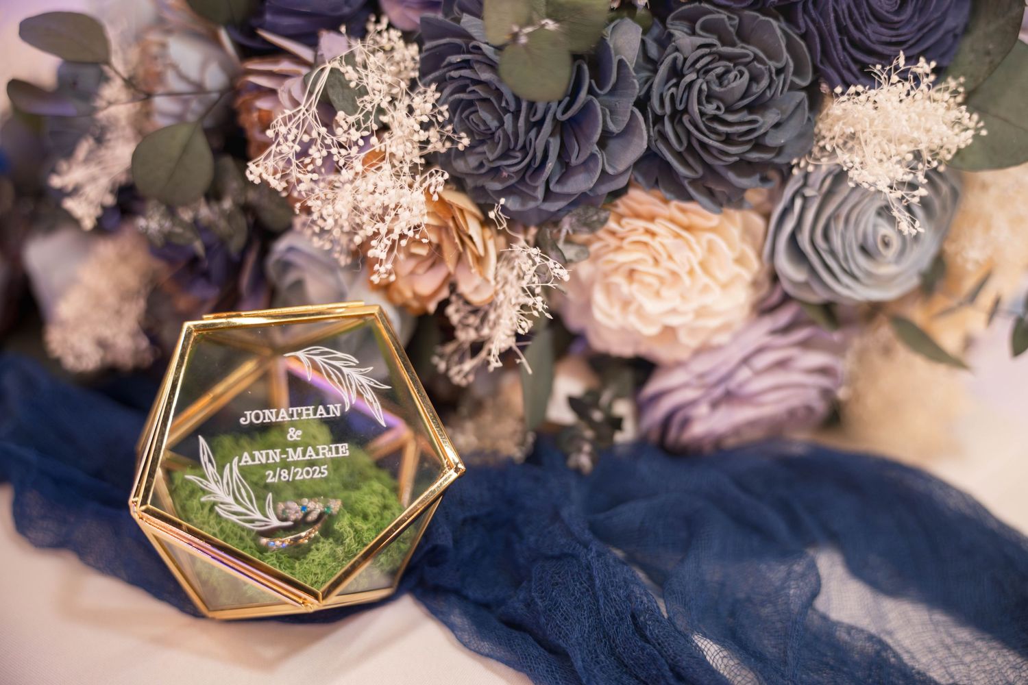 Glass hexagonal ring box decorated with floral accents and dried flowers on navy blue fabric.
