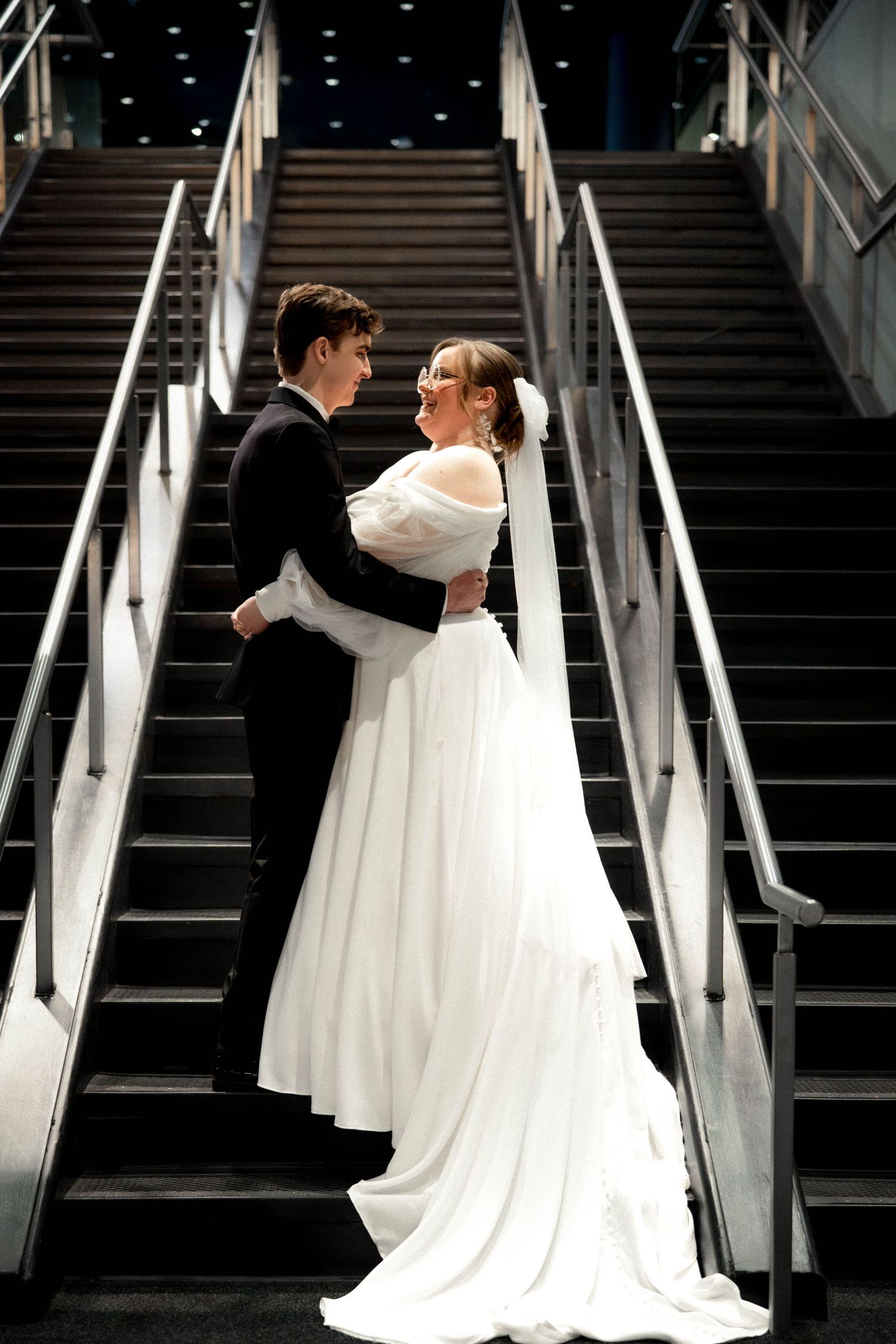 A couple in wedding attire stand together on a dramatic black staircase with metal railings.