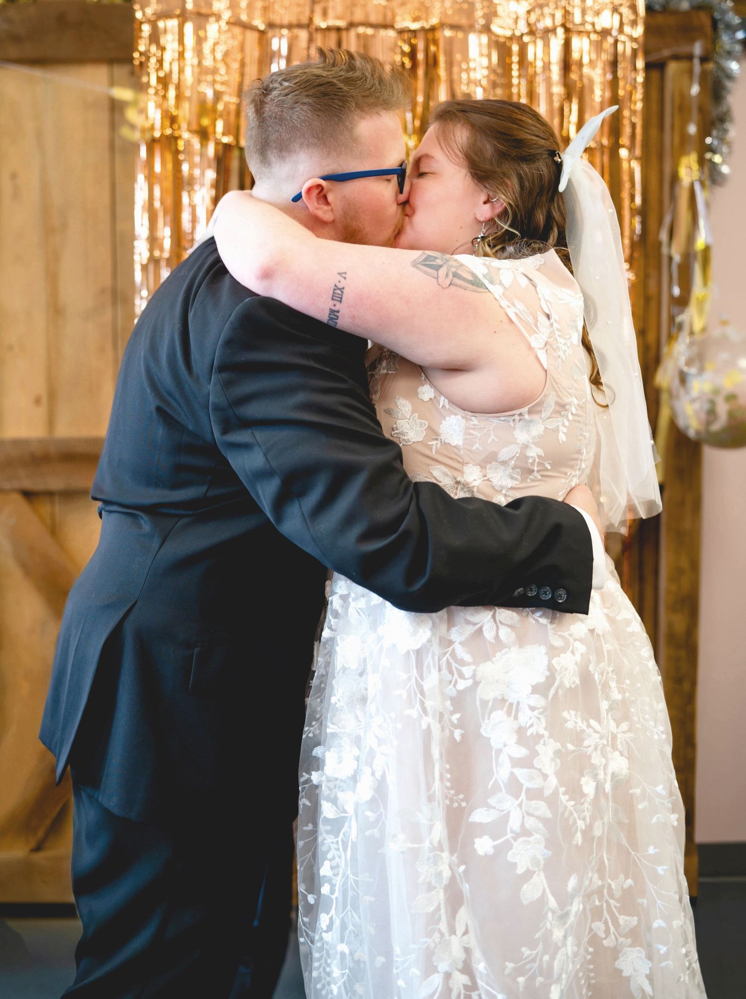 Two newlyweds share their first kiss during a wedding ceremony in front of a sparkly gold backdrop.
