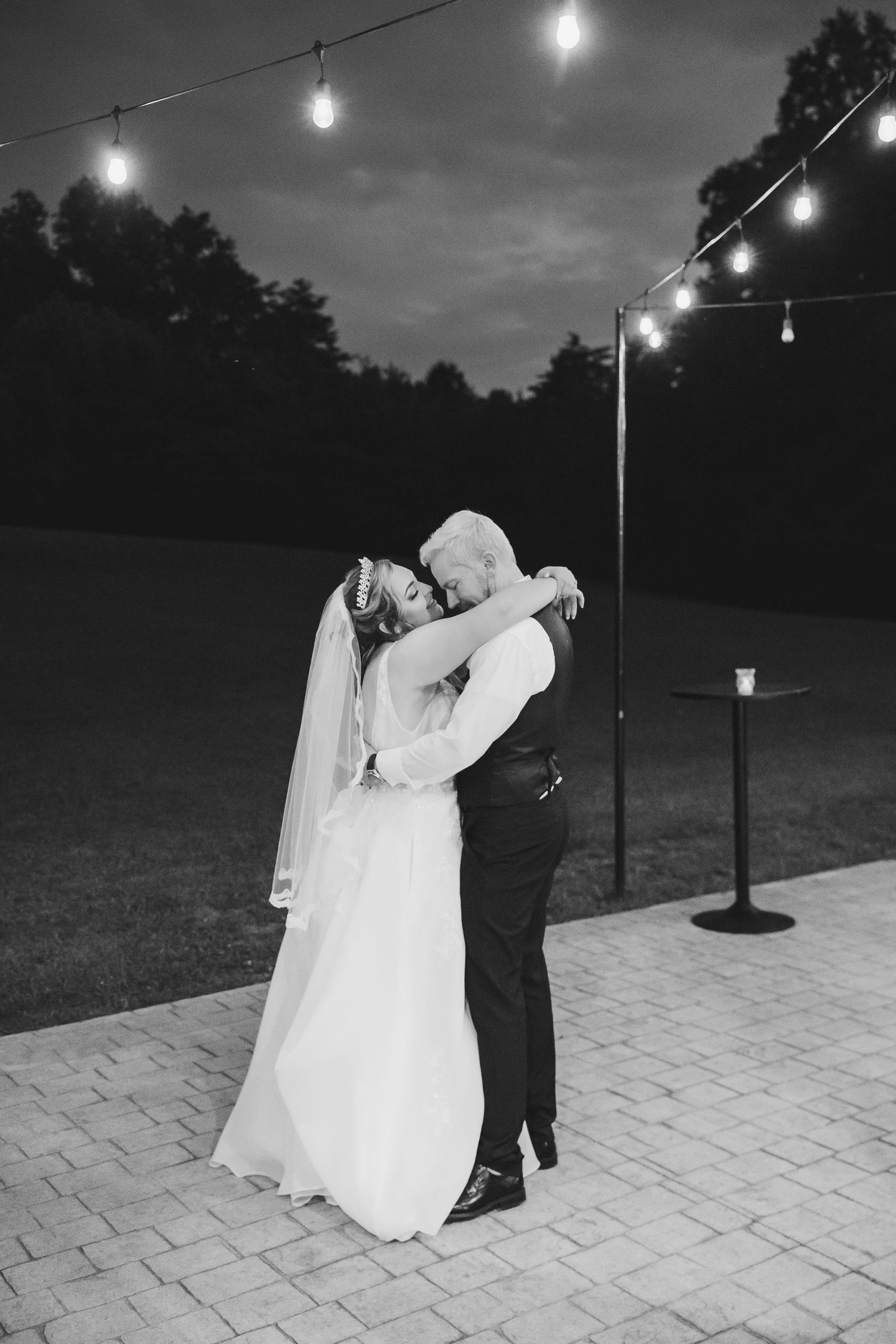 A couple shares a romantic dance under string lights on a stone patio at dusk.