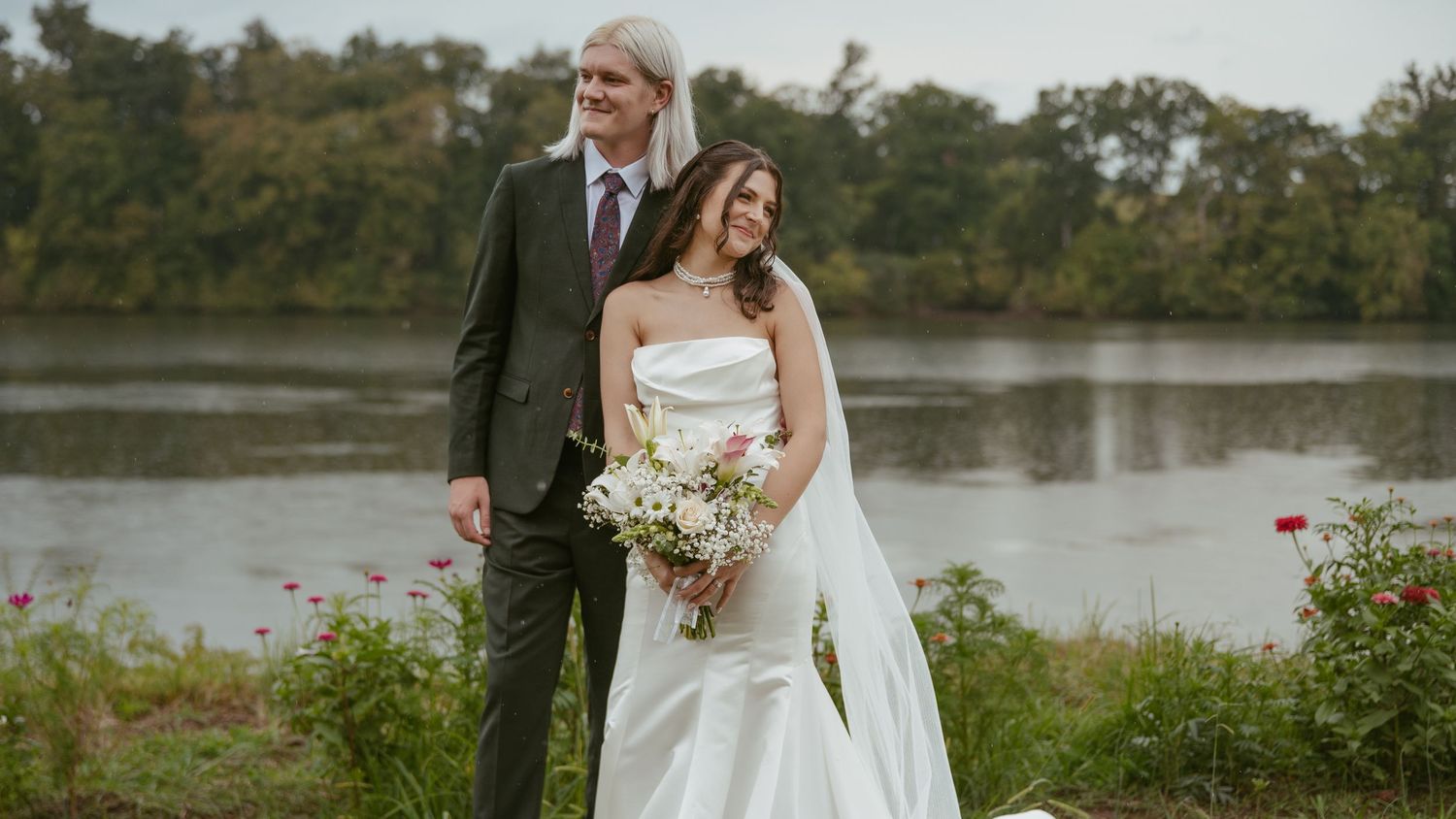 A newlywed couple stands together by a serene river in East Tennessee with flowers blooming in the foreground.