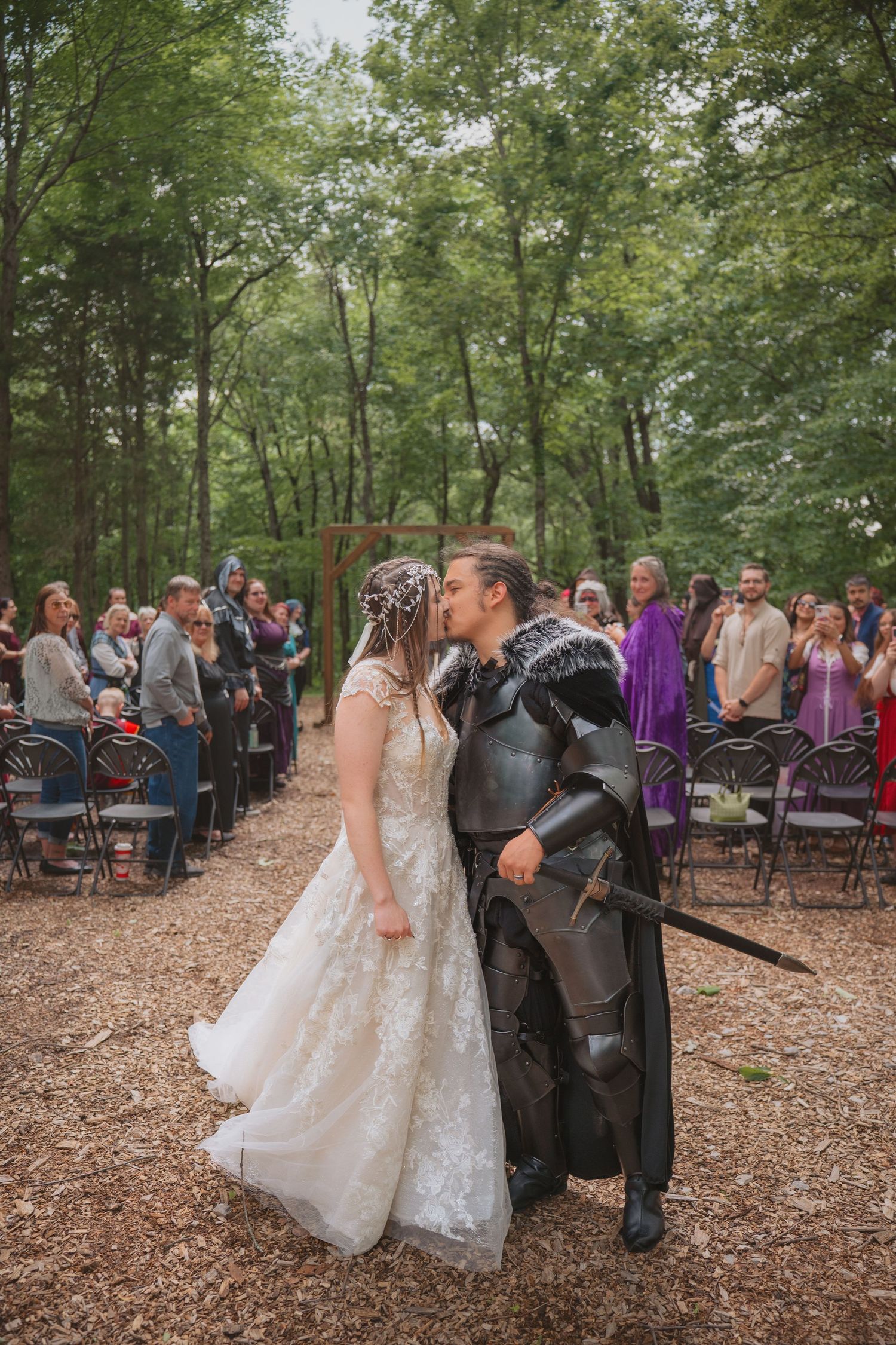 An outdoor woodland wedding ceremony takes place in a forest setting with chairs arranged among fallen autumn leaves.