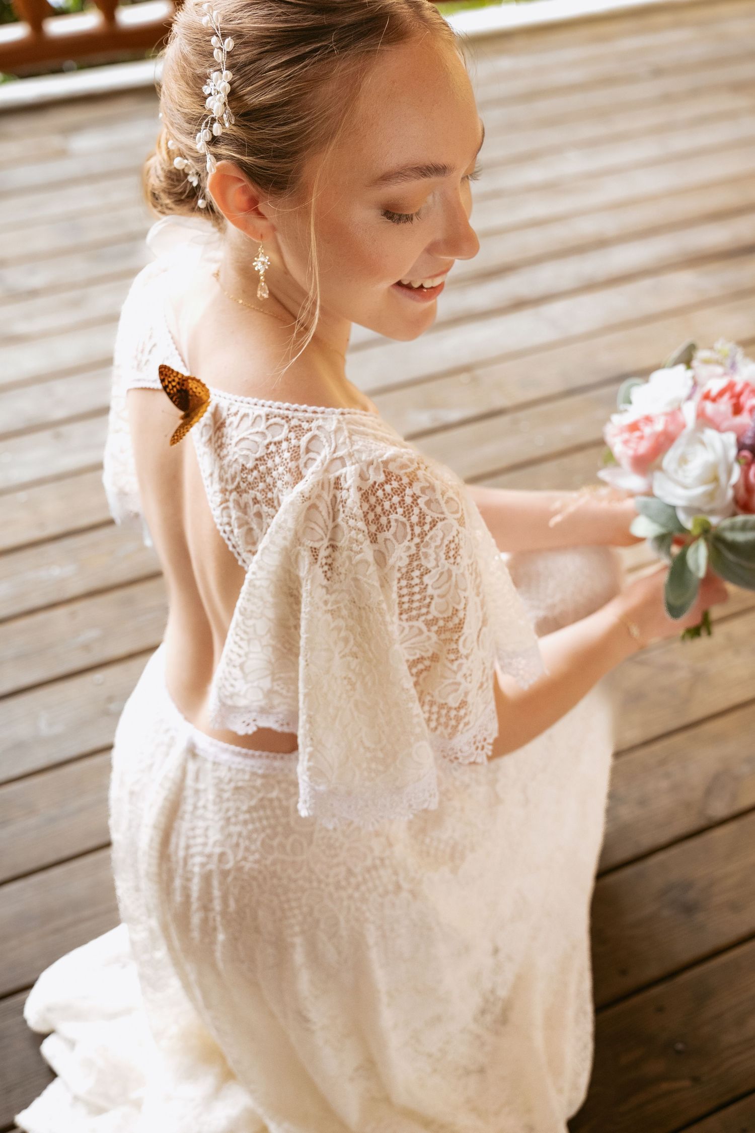 A bride in a delicate lace wedding dress poses on rustic wooden decking with soft natural lighting.