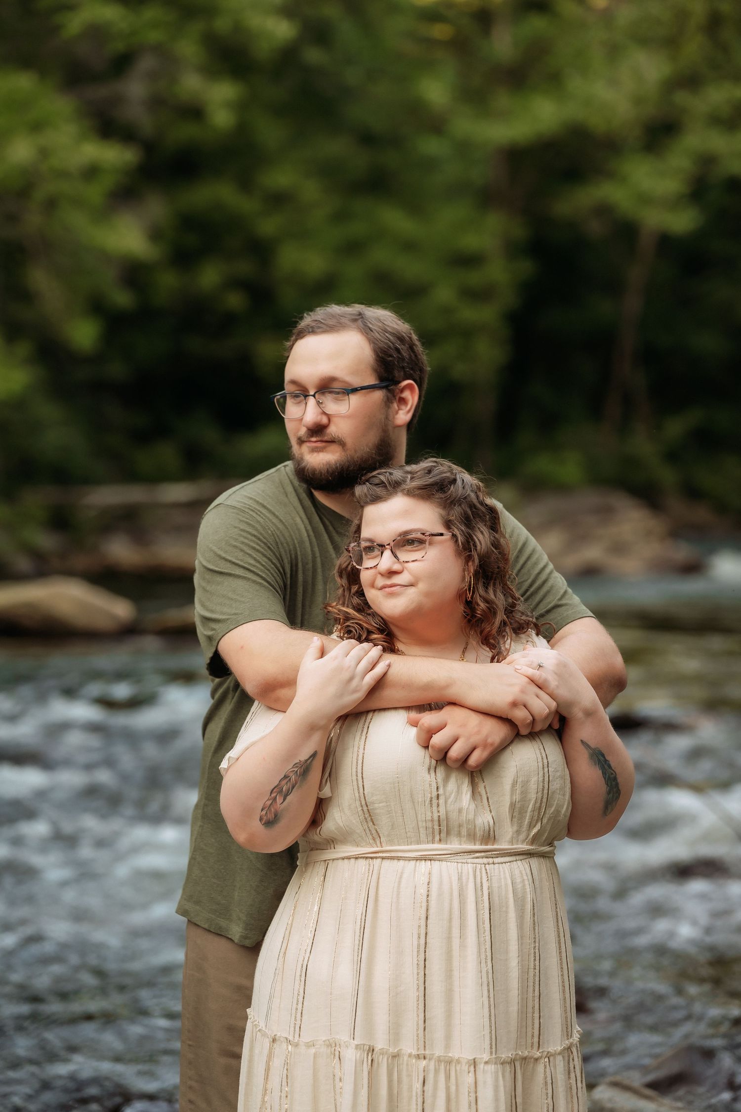 A couple embraces by a rushing river, wearing casual attire with glasses against a natural forest backdrop.