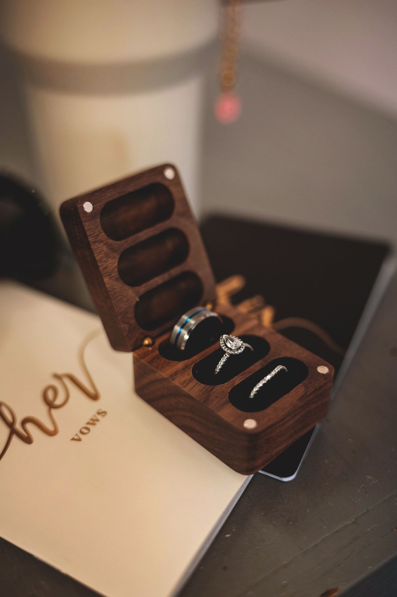 A wooden ring box with three wedding rings displayed inside sits next to a paper card on a dark surface.