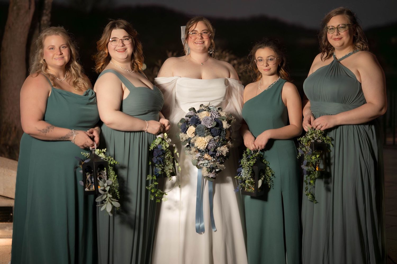 Wedding party in sage green bridesmaid dresses and white wedding gown holding floral bouquets at twilight.