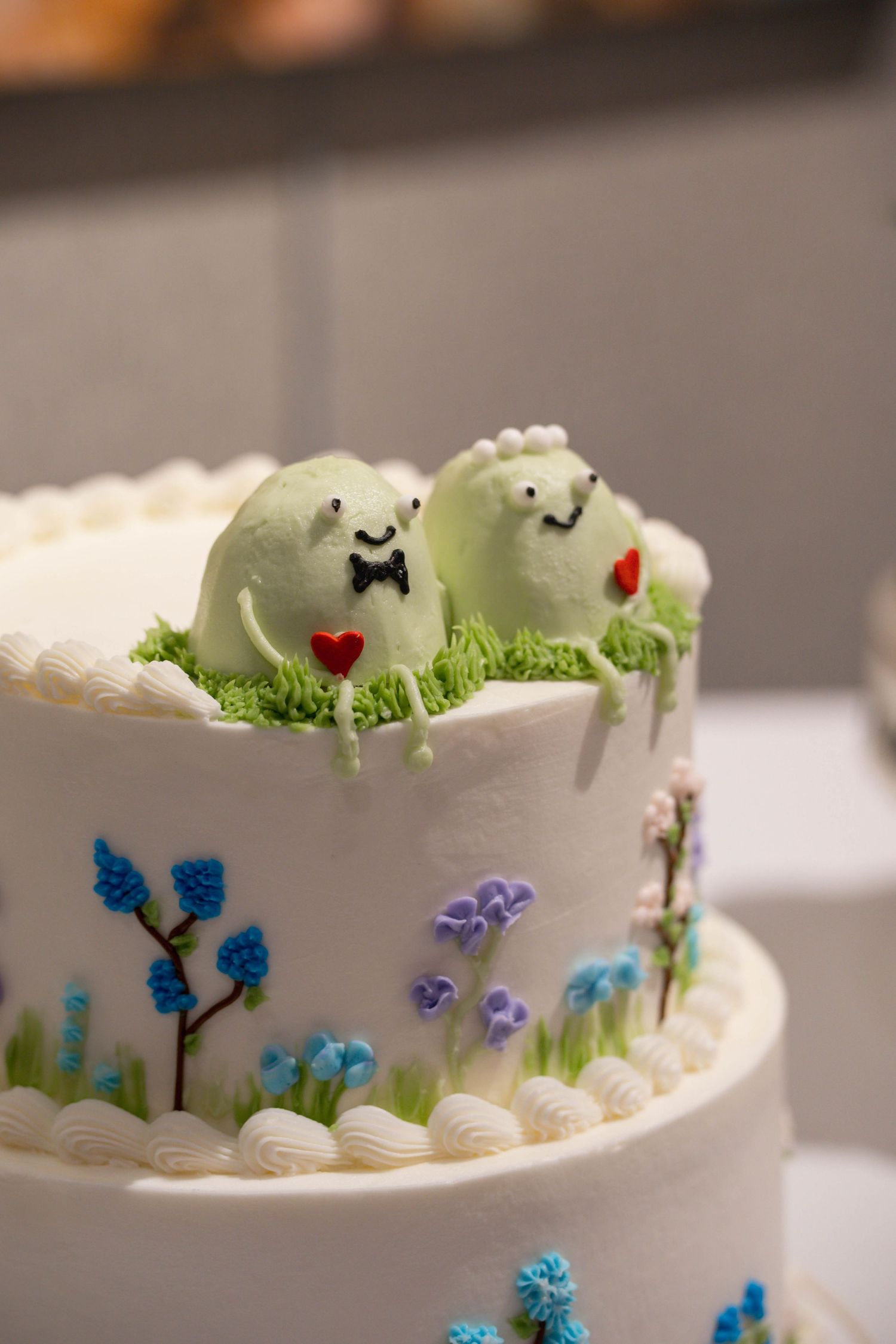 White tiered wedding cake decorated with miniature green dinosaur cake toppers and colorful floral designs on the sides.