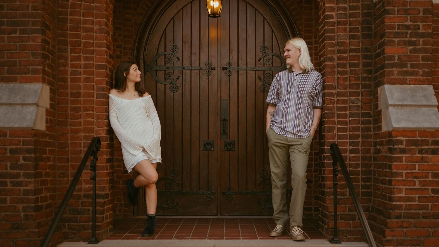 Two people stand casually in front of a large wooden door at a brick building entrance.