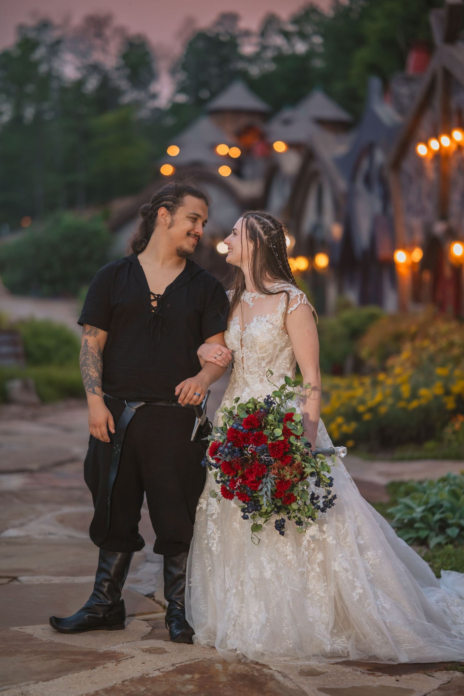 Couple in wedding attire share a moment at dusk with twinkling lights and a vibrant red bouquet.