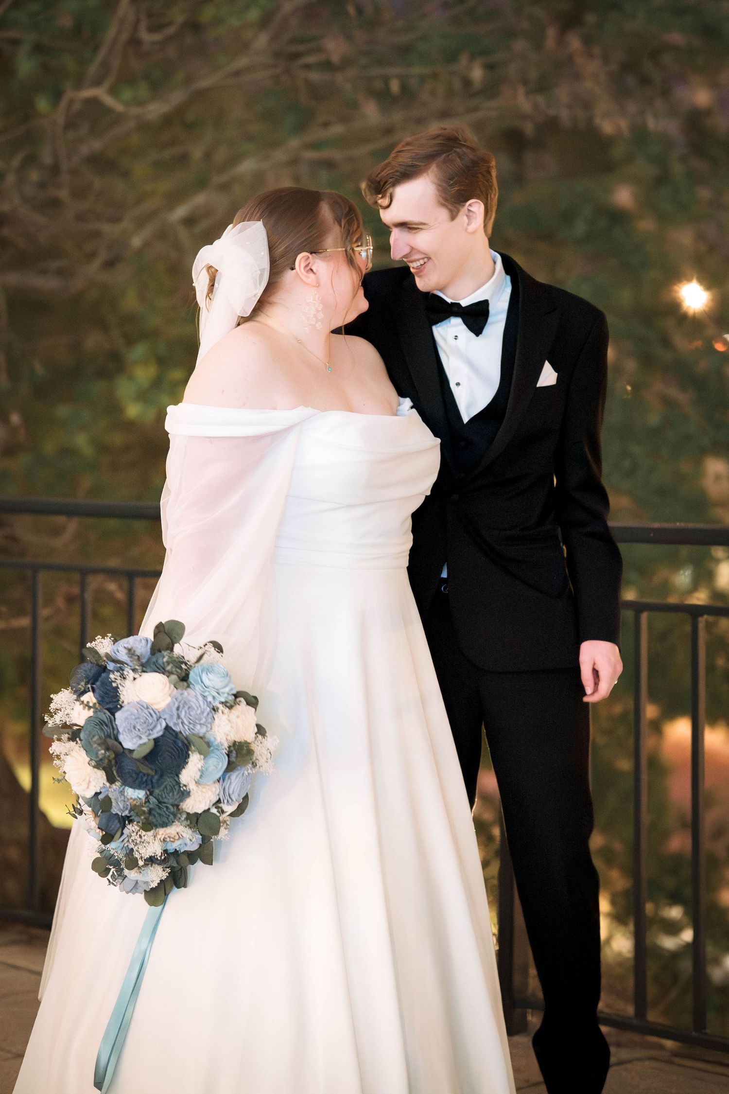 Newlywed couple shares an intimate moment on a balcony at dusk while bride holds a blue and white floral bouquet.