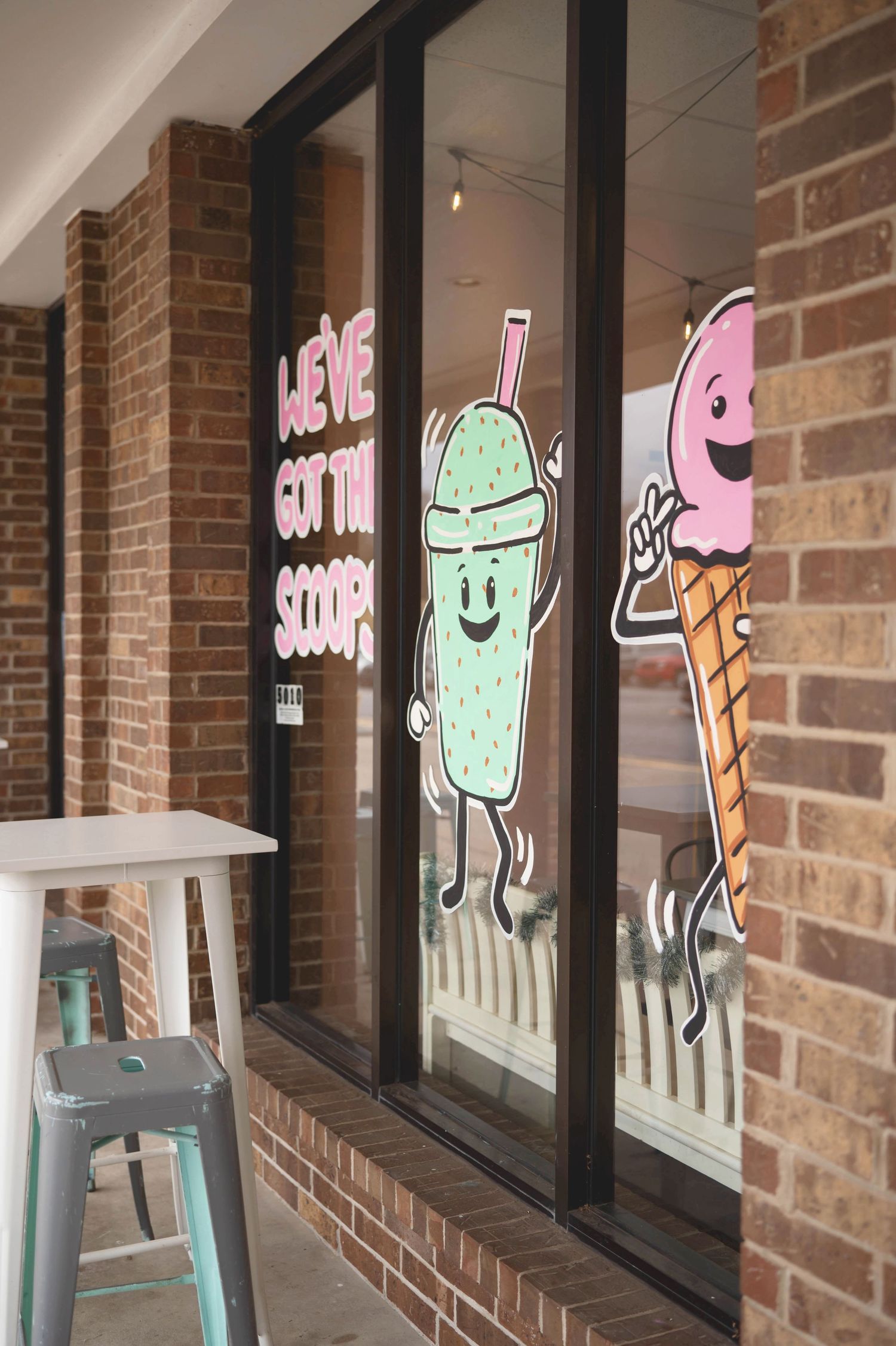 Storefront window decorated with cute cartoon illustrations of milkshake and ice cream characters on a brick building.
