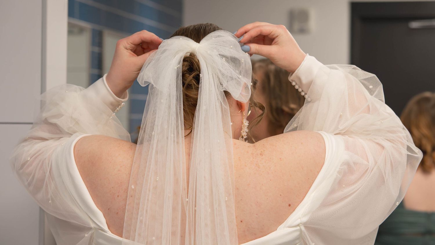 A wedding veil being arranged and pinned to the back of a white bridal gown with organza sleeves.