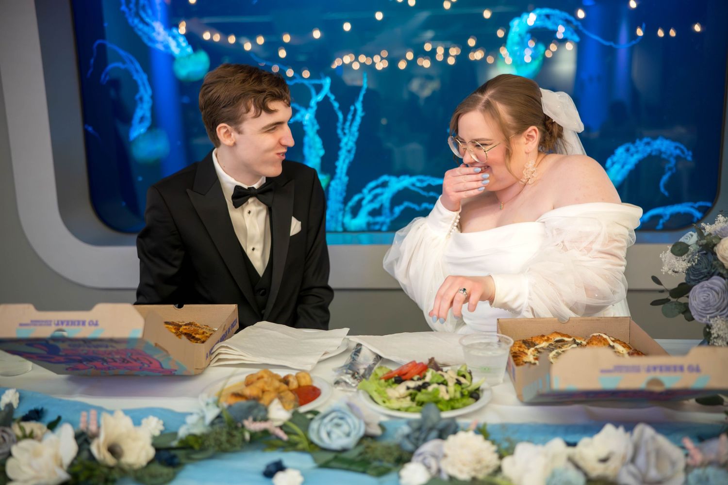 Wedding couple laughs together at their reception dinner surrounded by twinkle lights and floral decor.