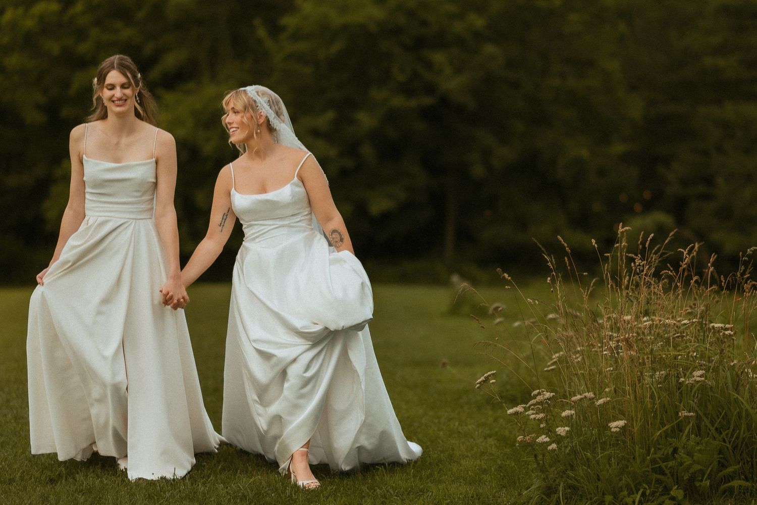 Two elegant white dresses move gracefully across a meadow with wildflowers at sunset.