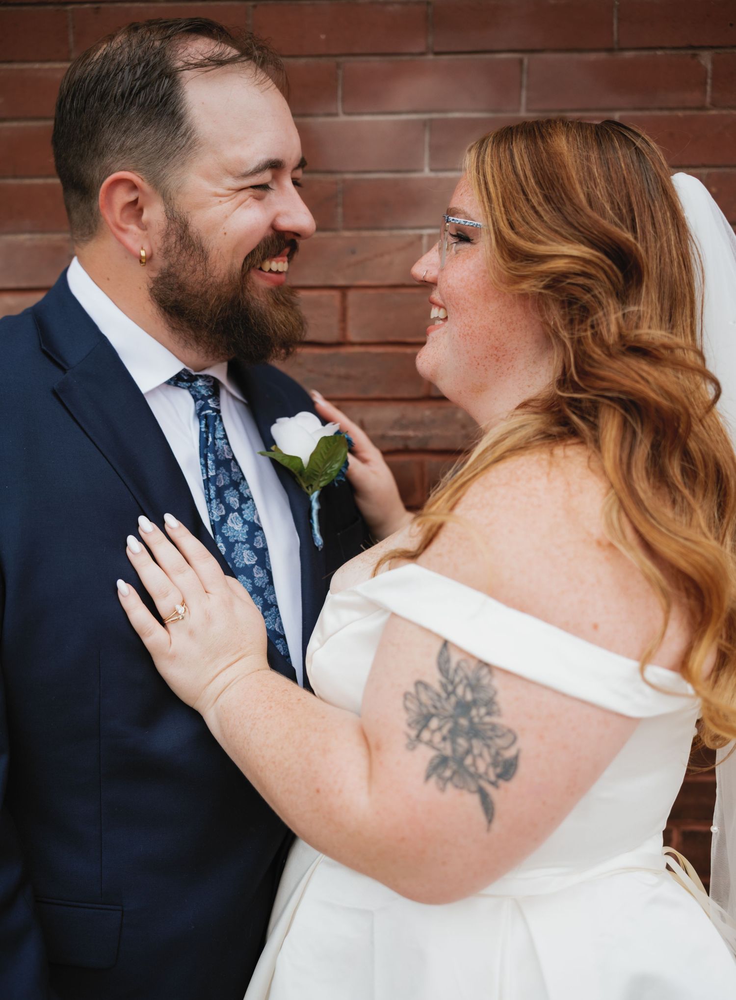 A couple dressed in wedding attire embrace and smile against a brick wall background.