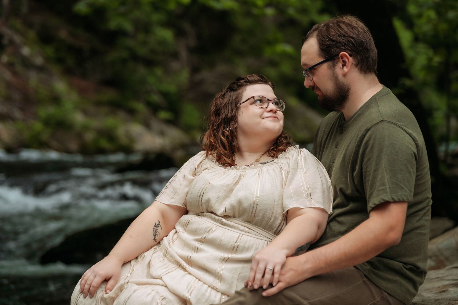 A romantic couple poses together by a river in a natural outdoor setting during an engagement photo session.