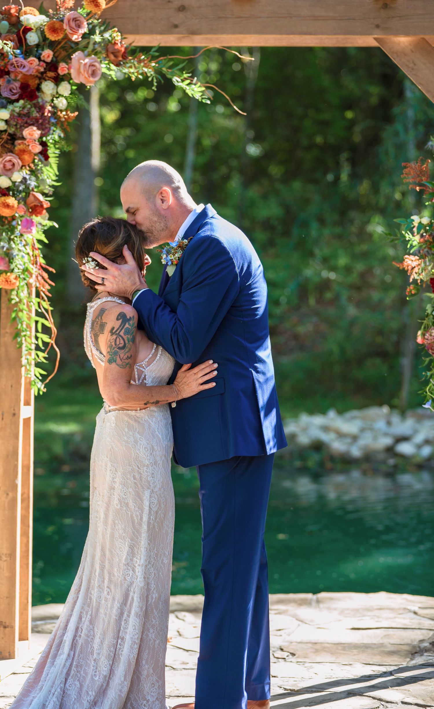 Couple sharing an intimate embrace under a wooden pergola decorated with pink and peach floral arrangements.