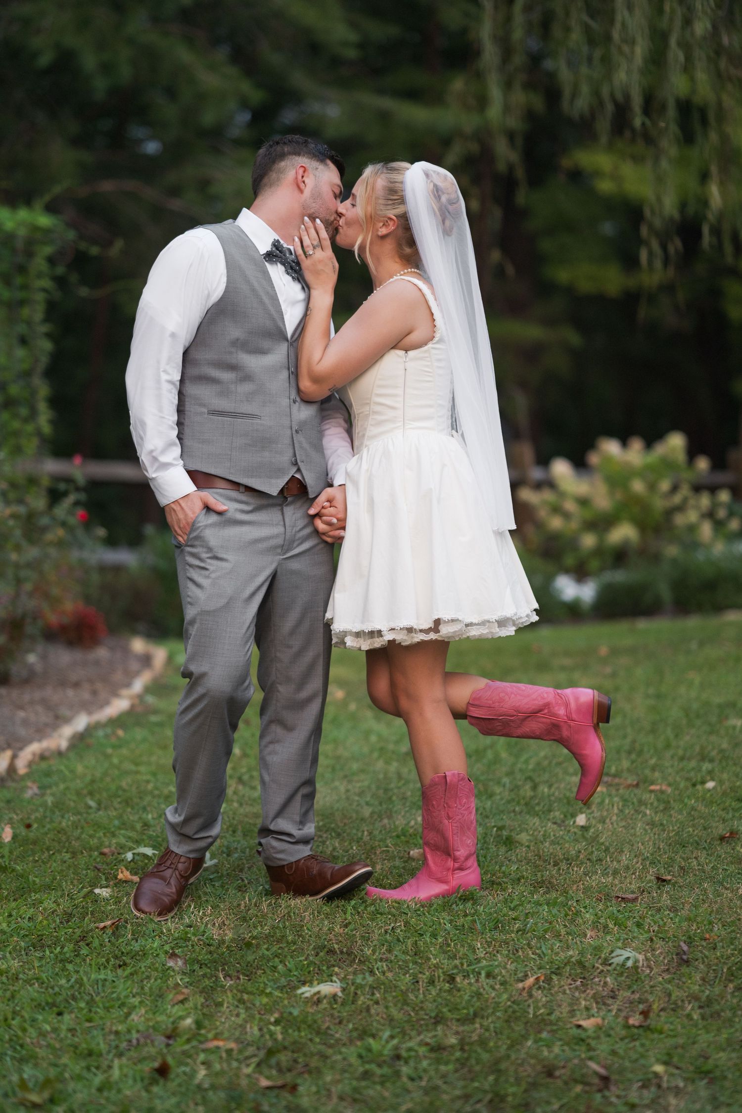 Couple shares romantic moment in garden, bride wearing short white dress and pink cowboy boots with groom in grey vest.