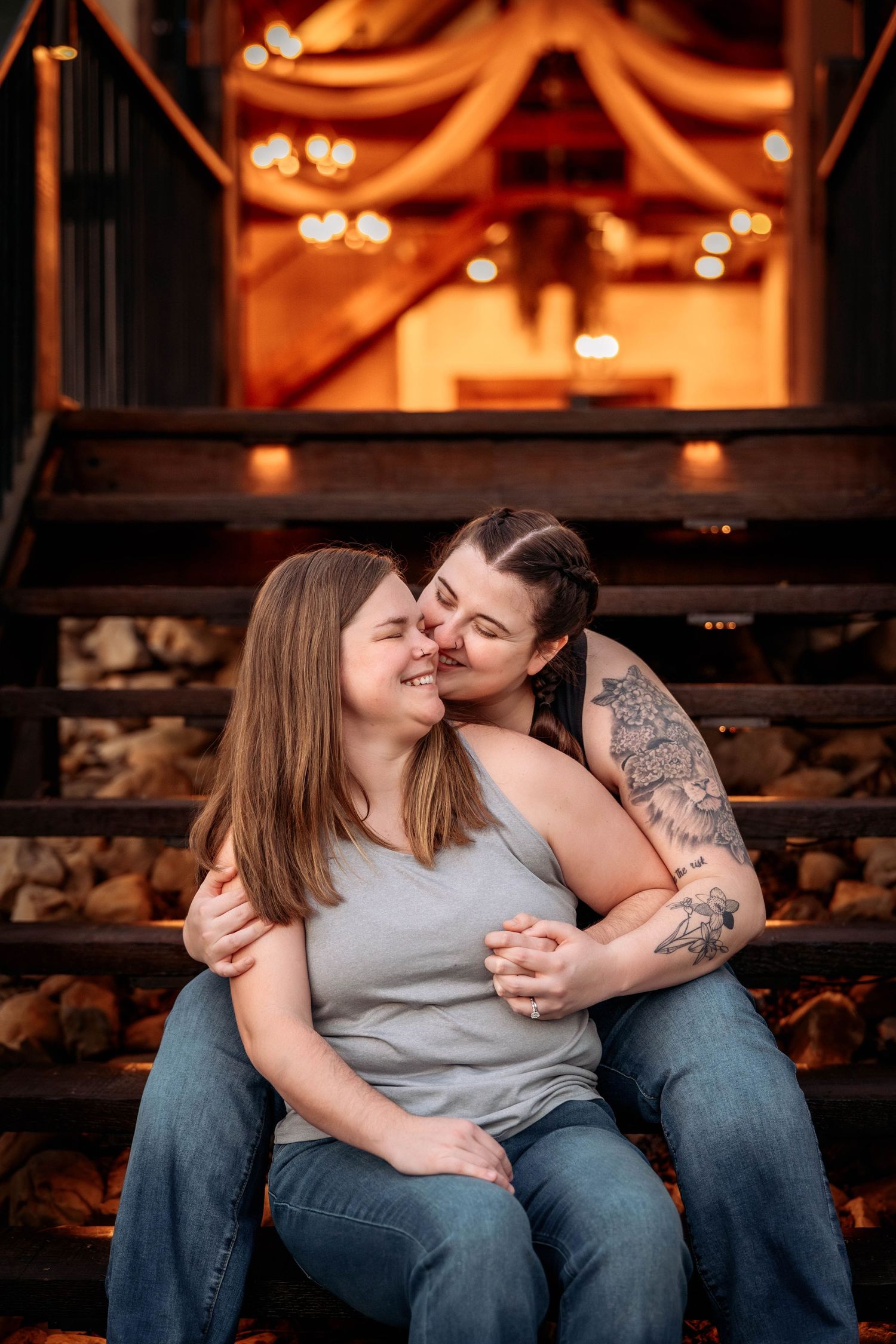 Romantic couple shares tender moment on wooden steps with warm glowing lights and rustic decor in background.