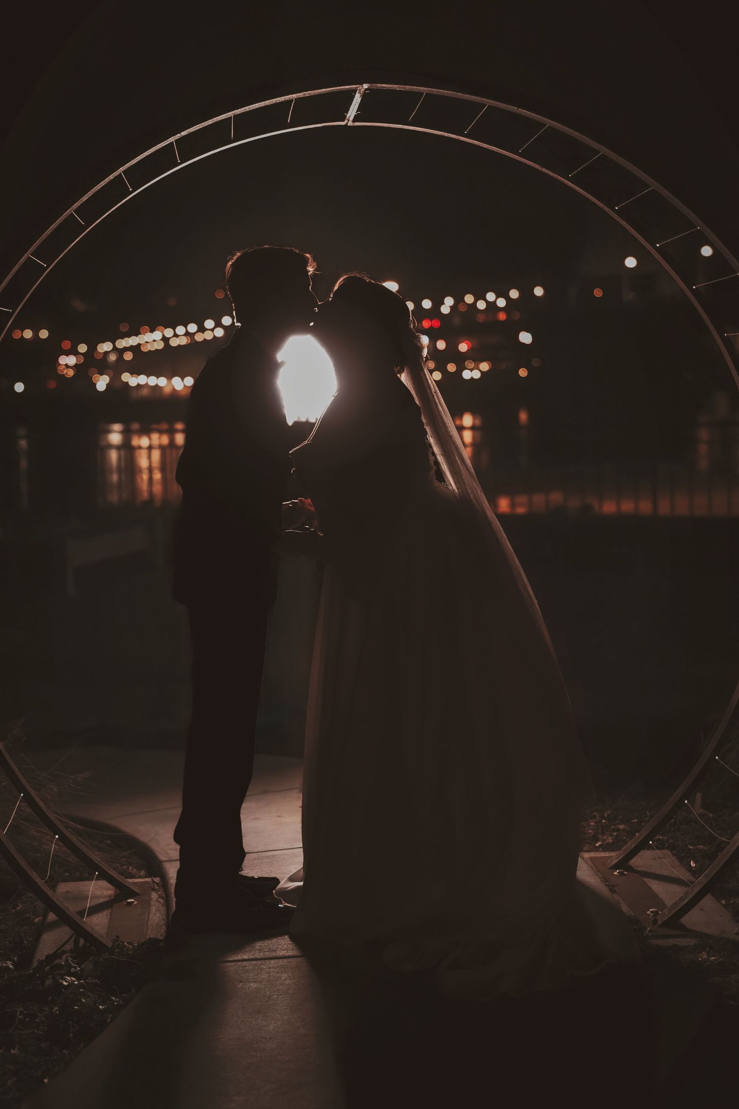A romantic silhouette of a couple standing in an archway at night with twinkling city lights in the background.