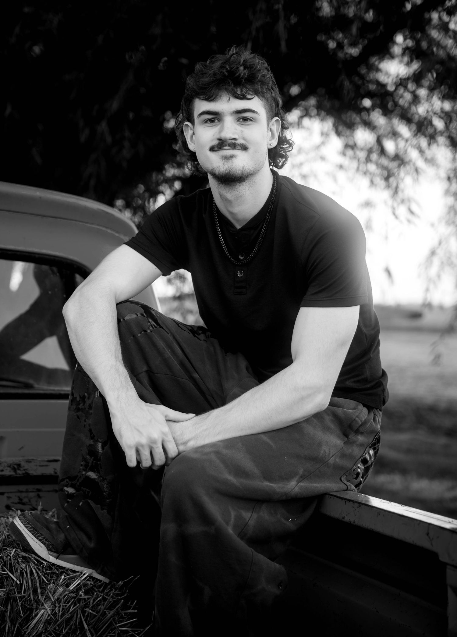 Black and white portrait of a smiling young man with curly hair and a mustache sitting on the tailgate of an old pickup truck outdoors.