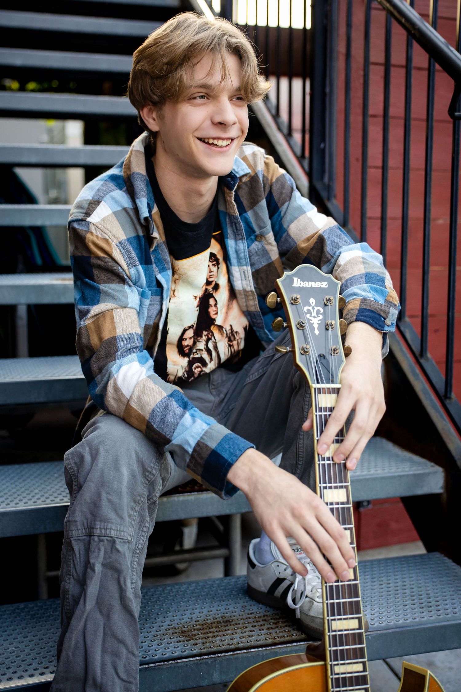 Smiling teen boy with guitar sitting on metal stairs wearing plaid shirt and band t-shirt.