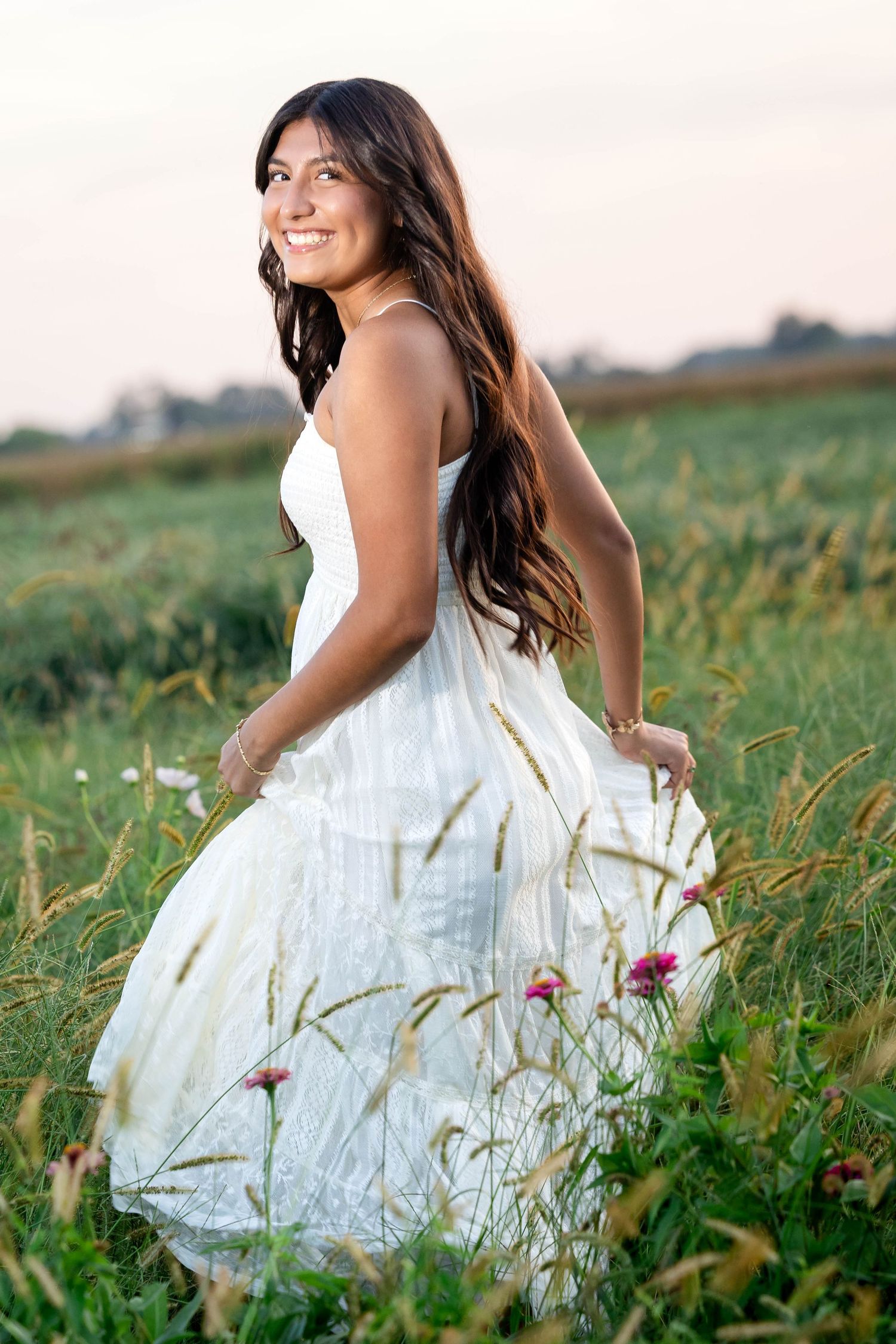 Smiling young woman with long dark hair in a flowing white sundress walks through a green meadow of wildflowers and tall grass at sunset.