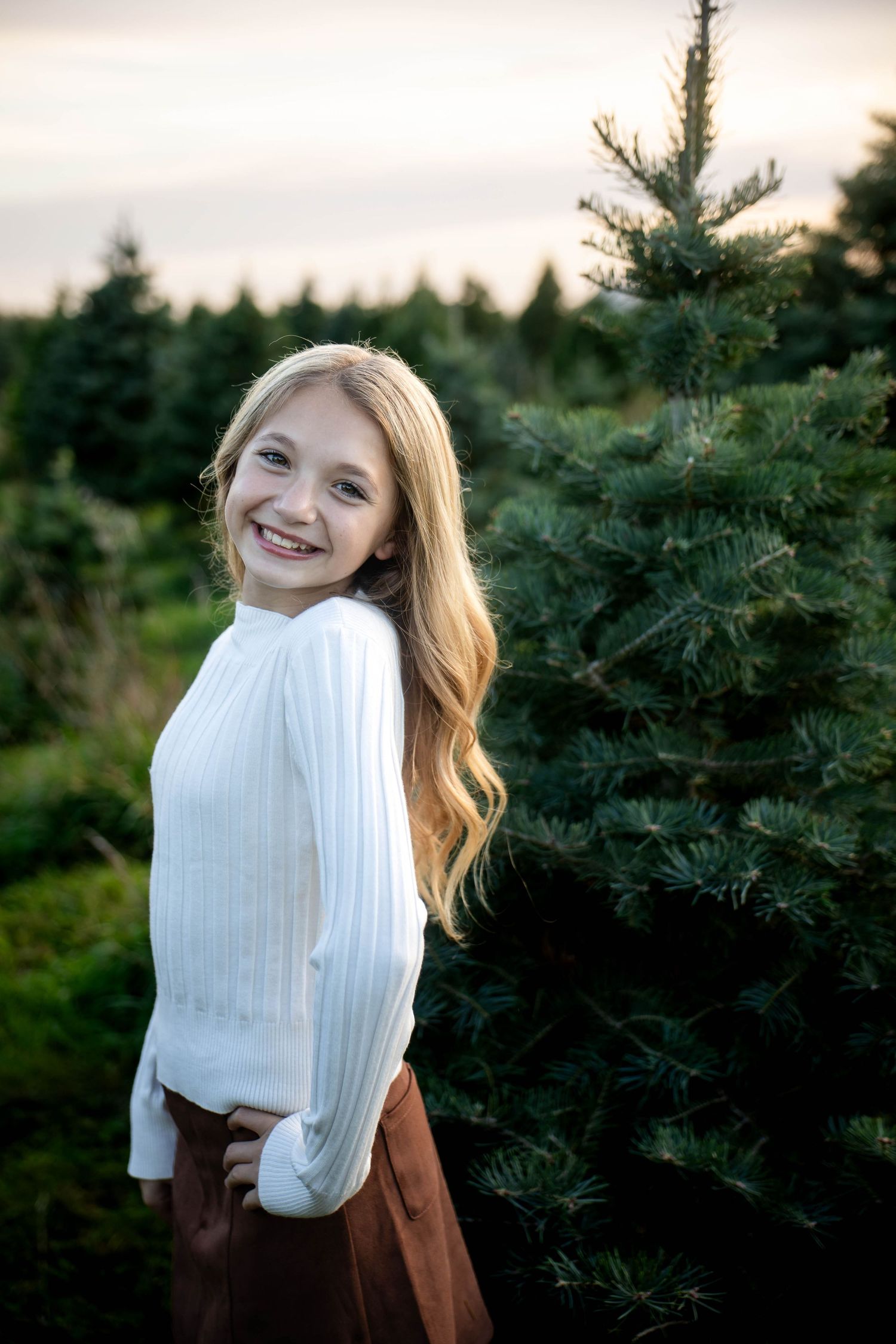 Smiling girl with long blonde hair in a white ribbed sweater and brown skirt poses among evergreen trees at a Christmas tree farm at sunset.