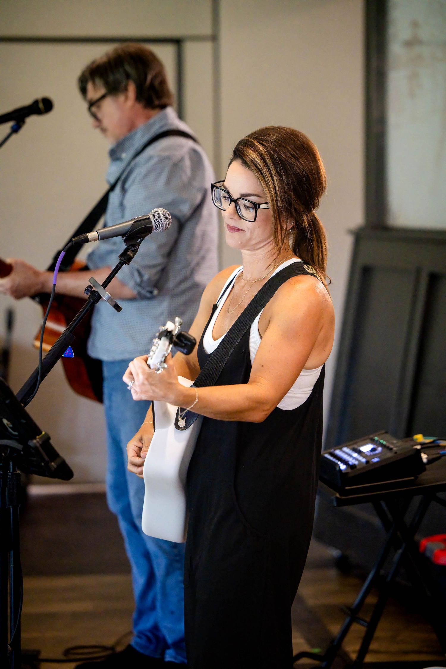 Female guitarist in black and white outfit plays guitar at an indoor music performance with bandmate behind her.