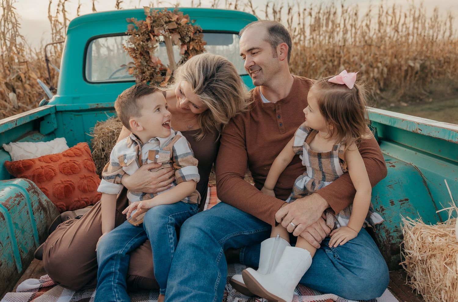 Happy family of four shares a joyful moment sitting in a vintage turquoise truck bed in a fall cornfield.