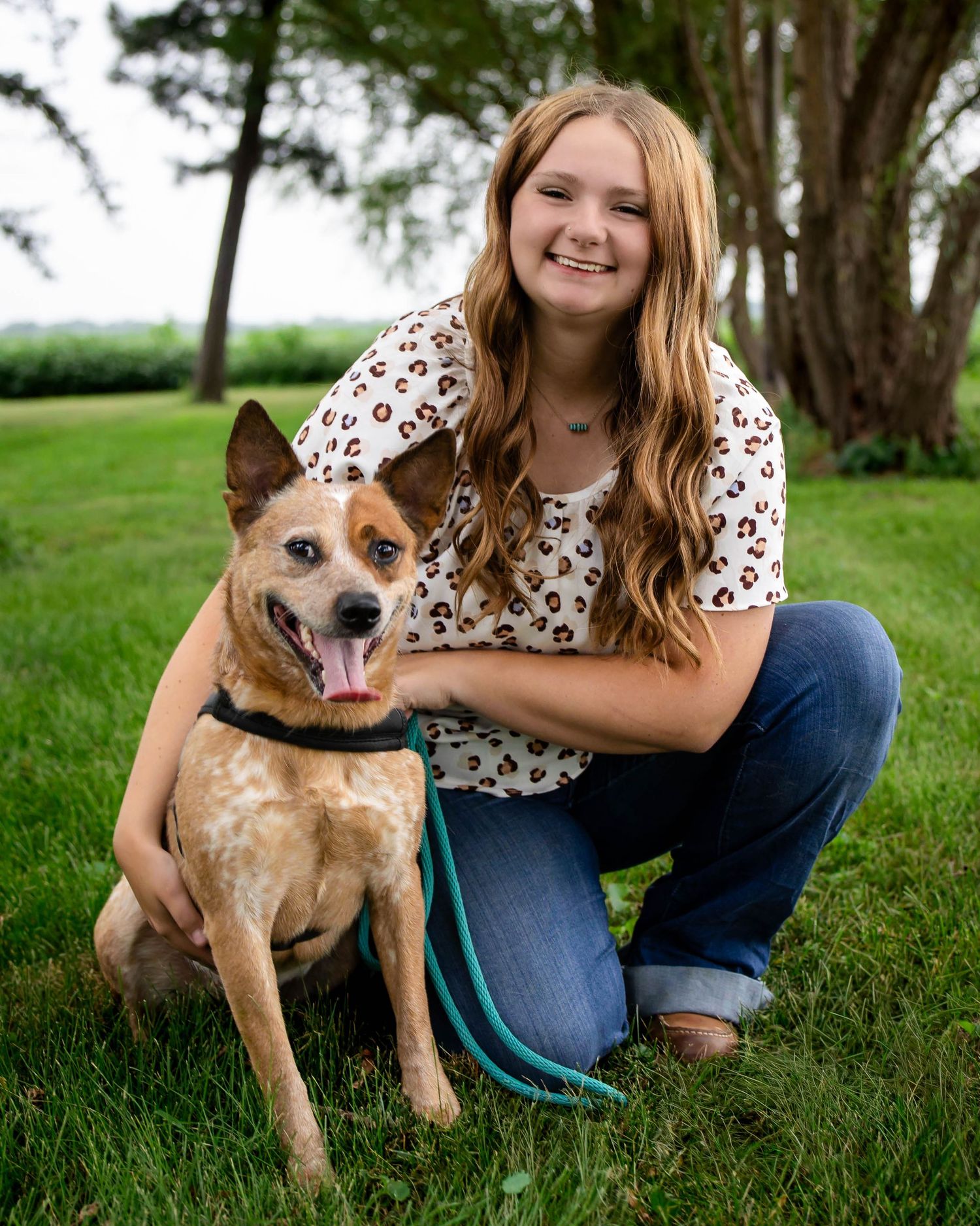 Smiling teenage girl crouching on grass with her red heeler dog on a leash outdoors.