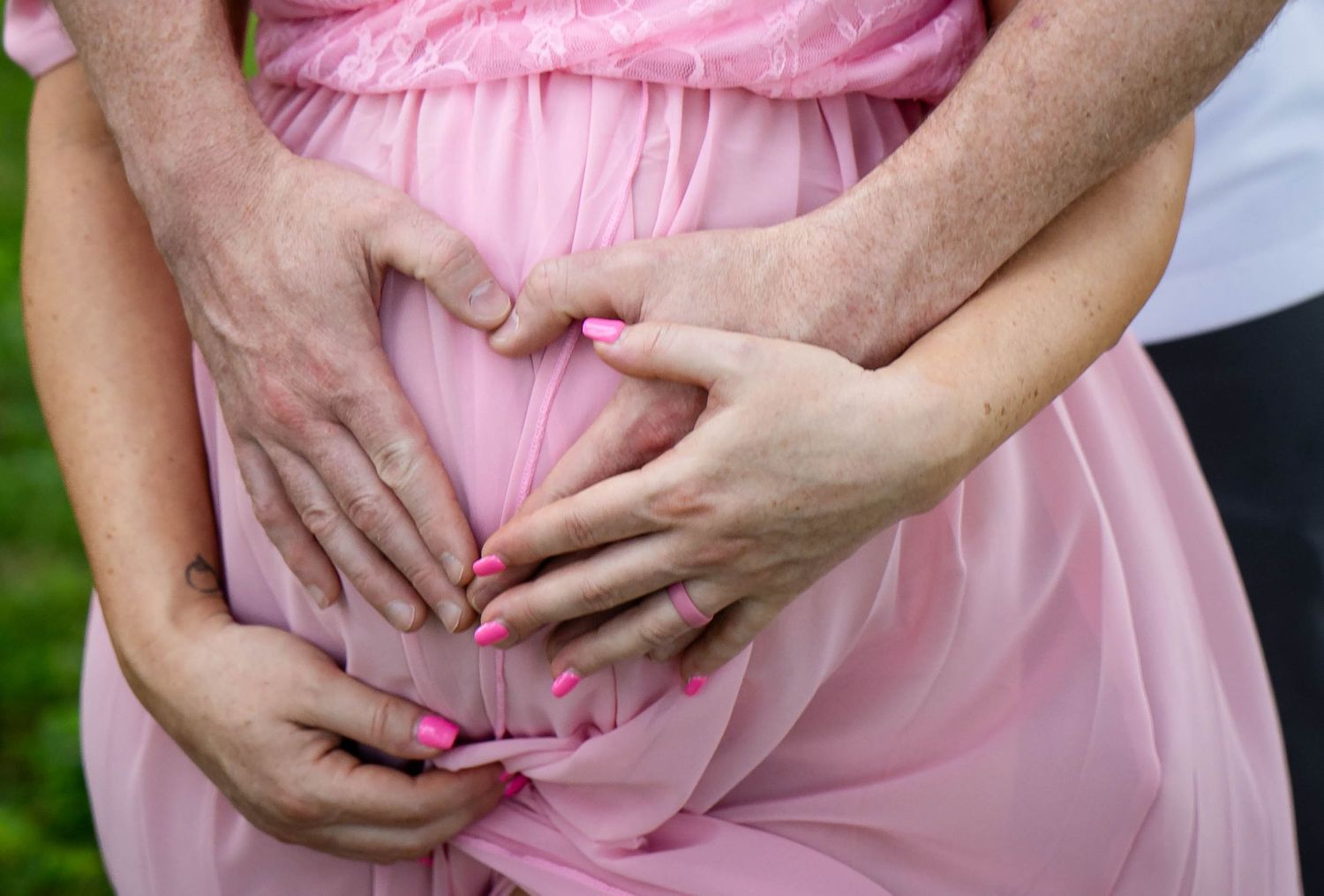 Pregnant woman in pink dress with family hands forming heart shape over baby bump outdoors.