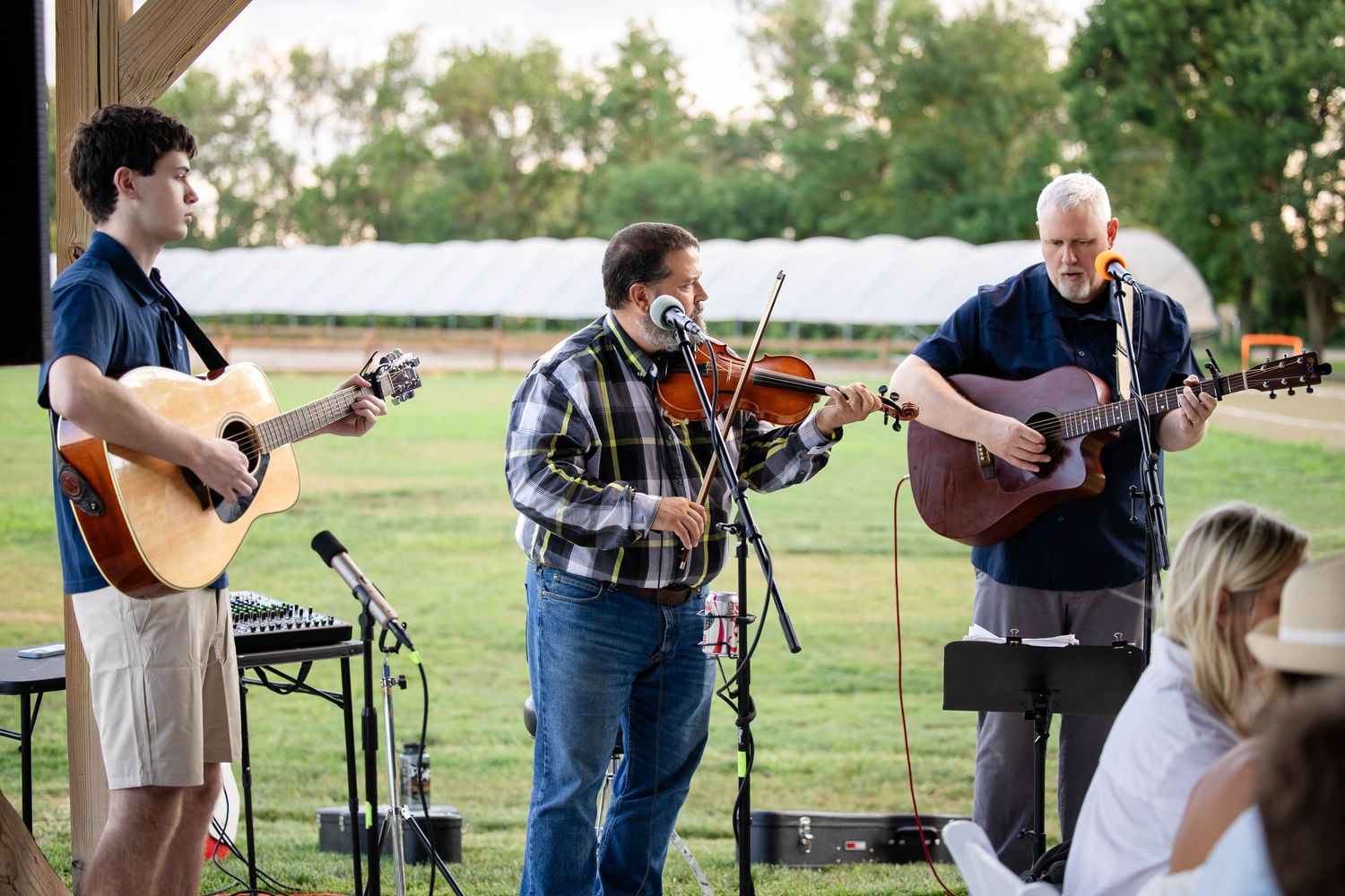 Three musicians perform bluegrass outdoors, playing violin and acoustic guitars on a rustic stage with green fields behind.