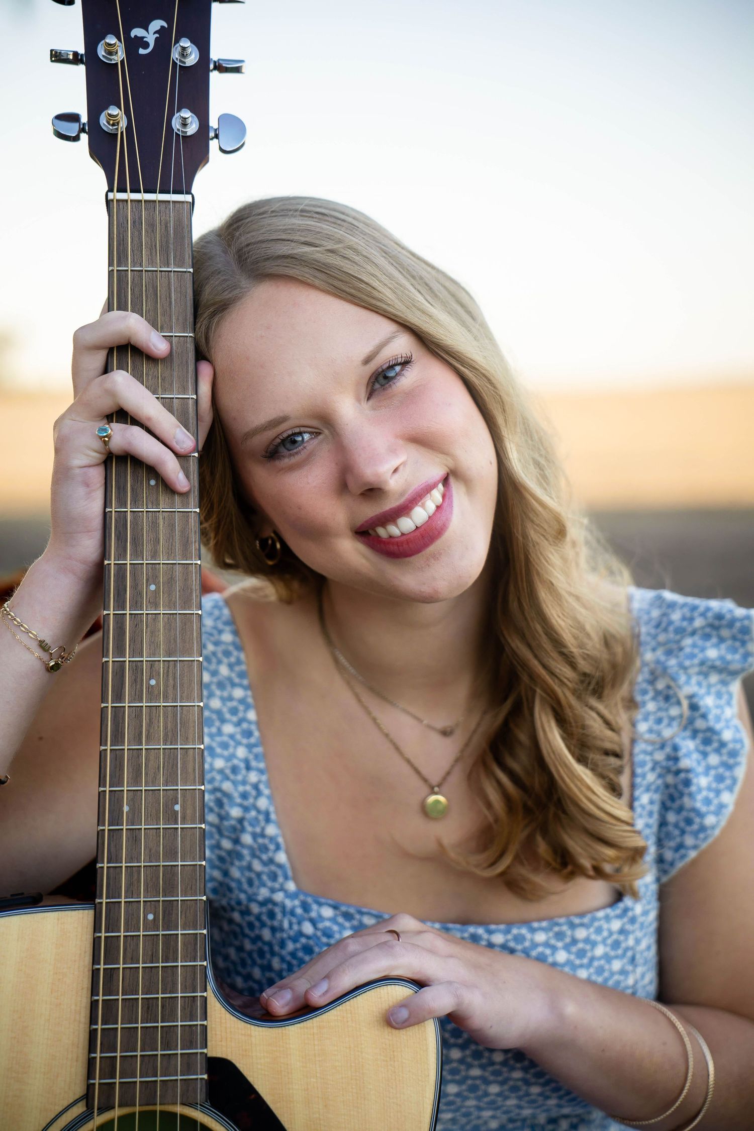 Smiling young woman with long blonde hair rests her head against the neck of an acoustic guitar while posing outdoors in a blue floral top.