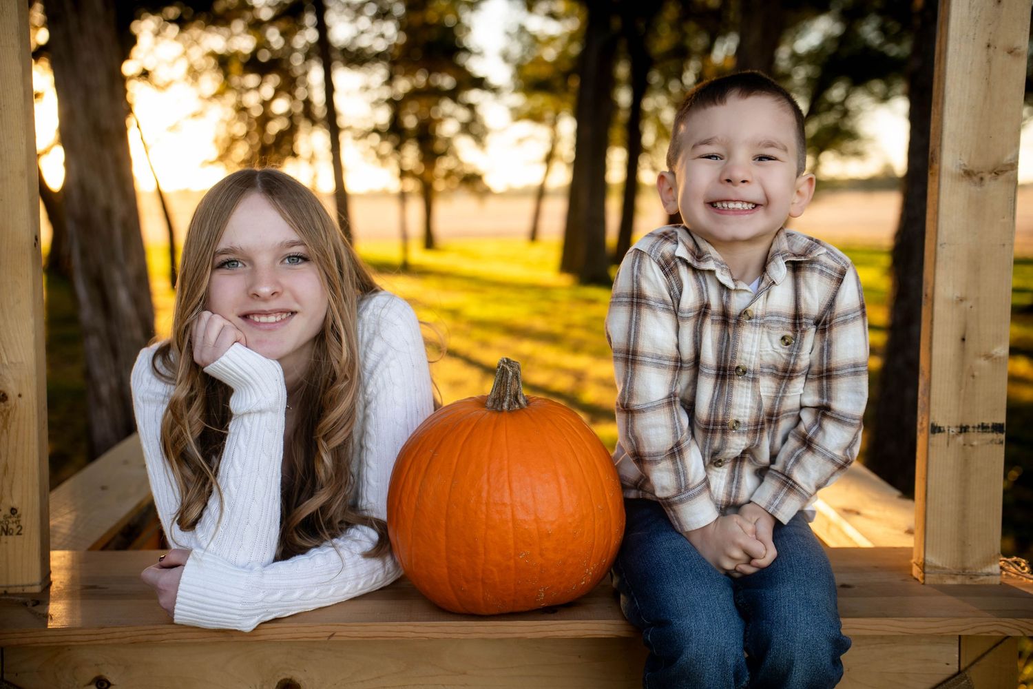 Smiling girl and boy sit on a wooden bench beside a large orange pumpkin in a sunlit autumn park with trees.