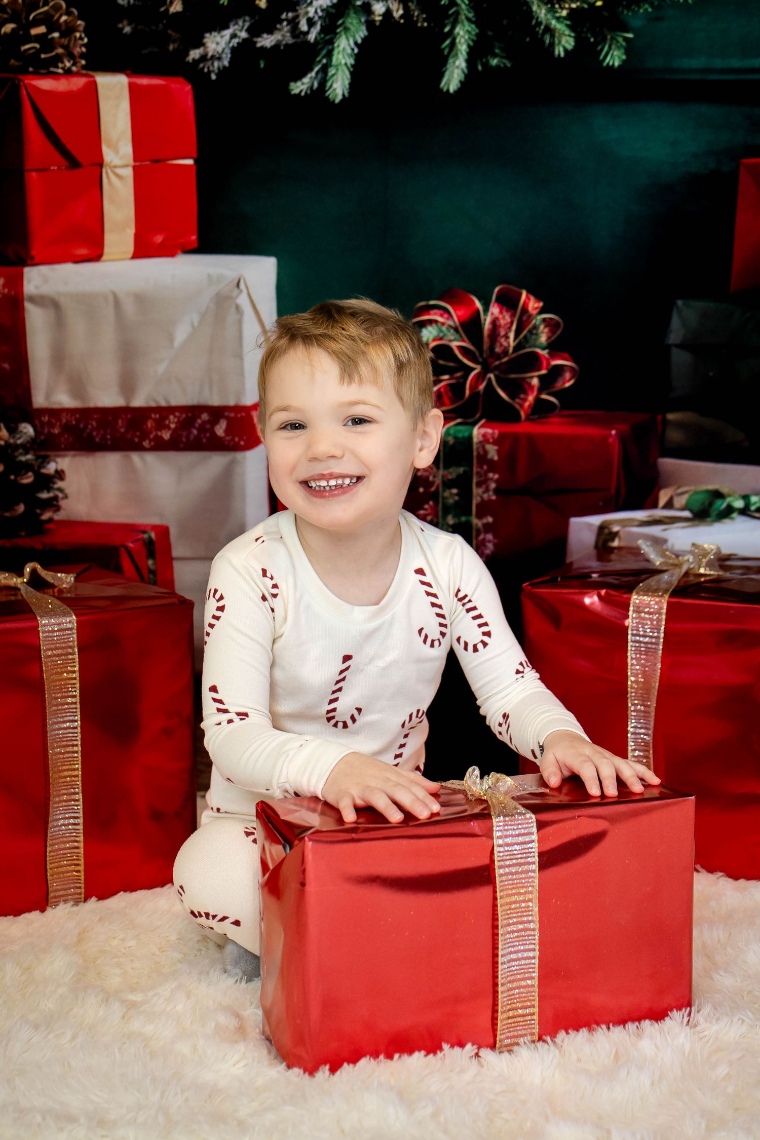 Smiling toddler in candy cane pajamas sits on a white fluffy rug with red Christmas gift boxes and festive bows behind him.