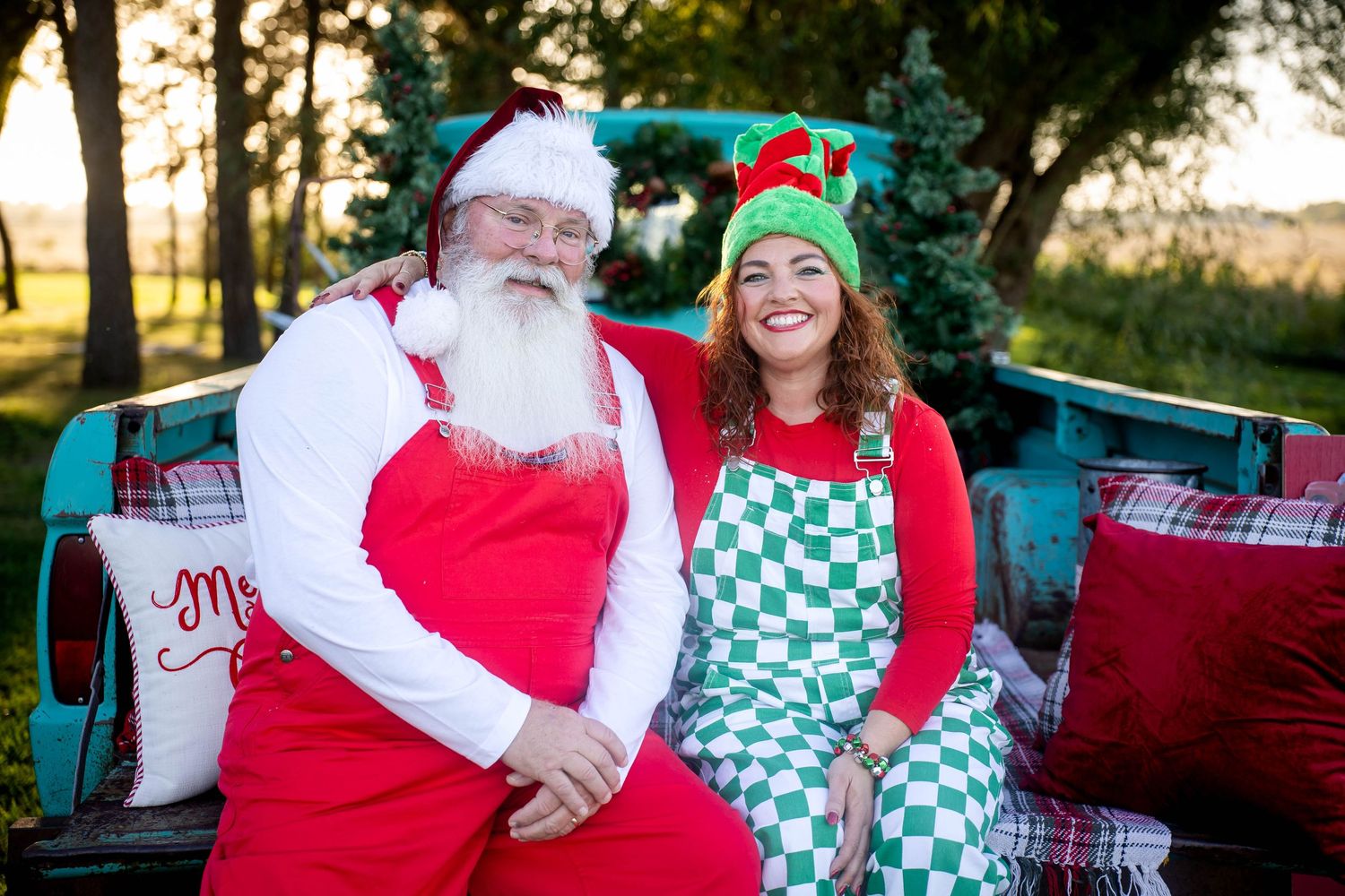 Santa Claus and a smiling woman in an elf hat and checkered apron sit together outdoors with Christmas decorations.