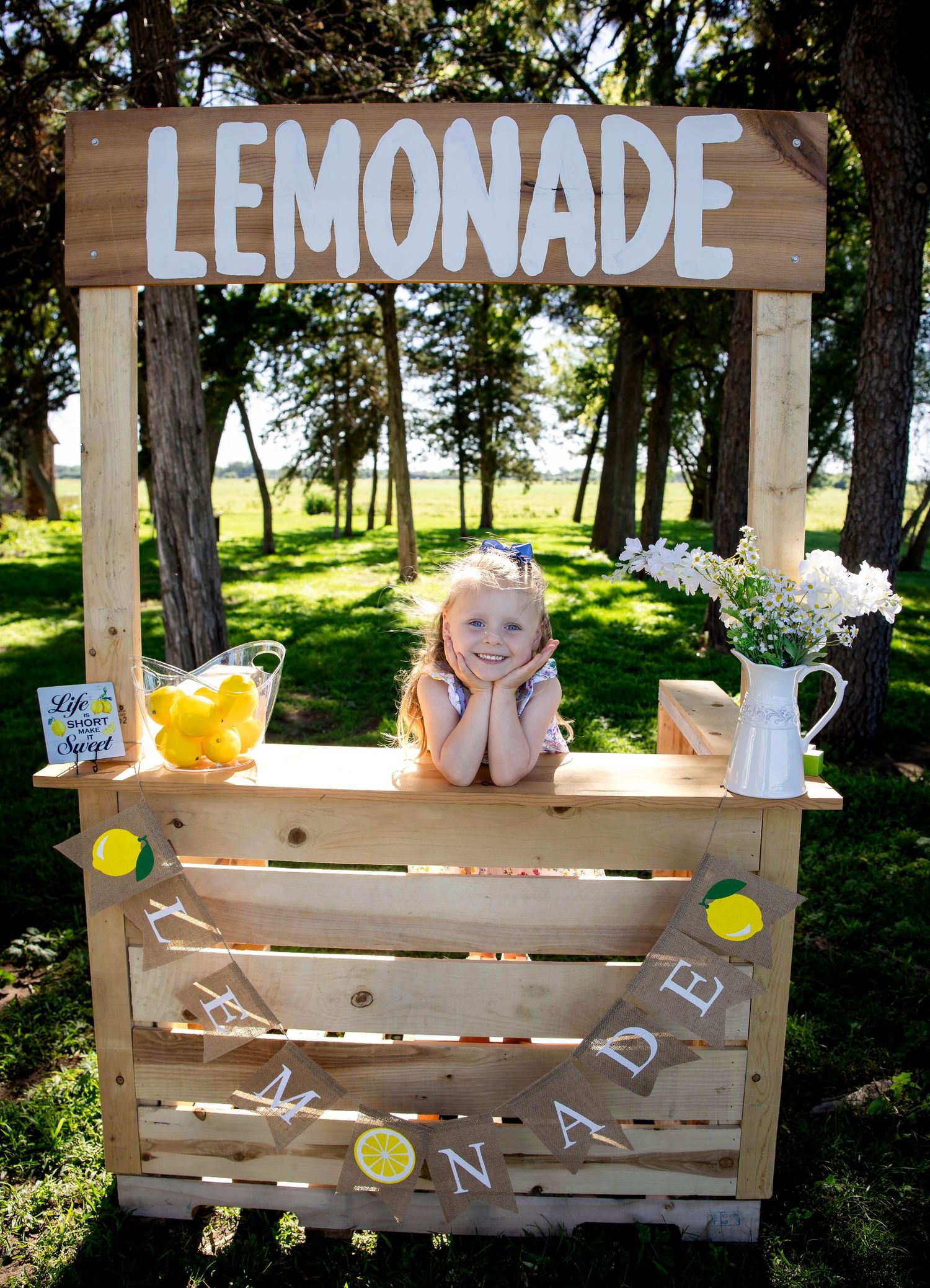 Adorable little girl smiling behind rustic wooden lemonade stand outdoors in grassy park setting.